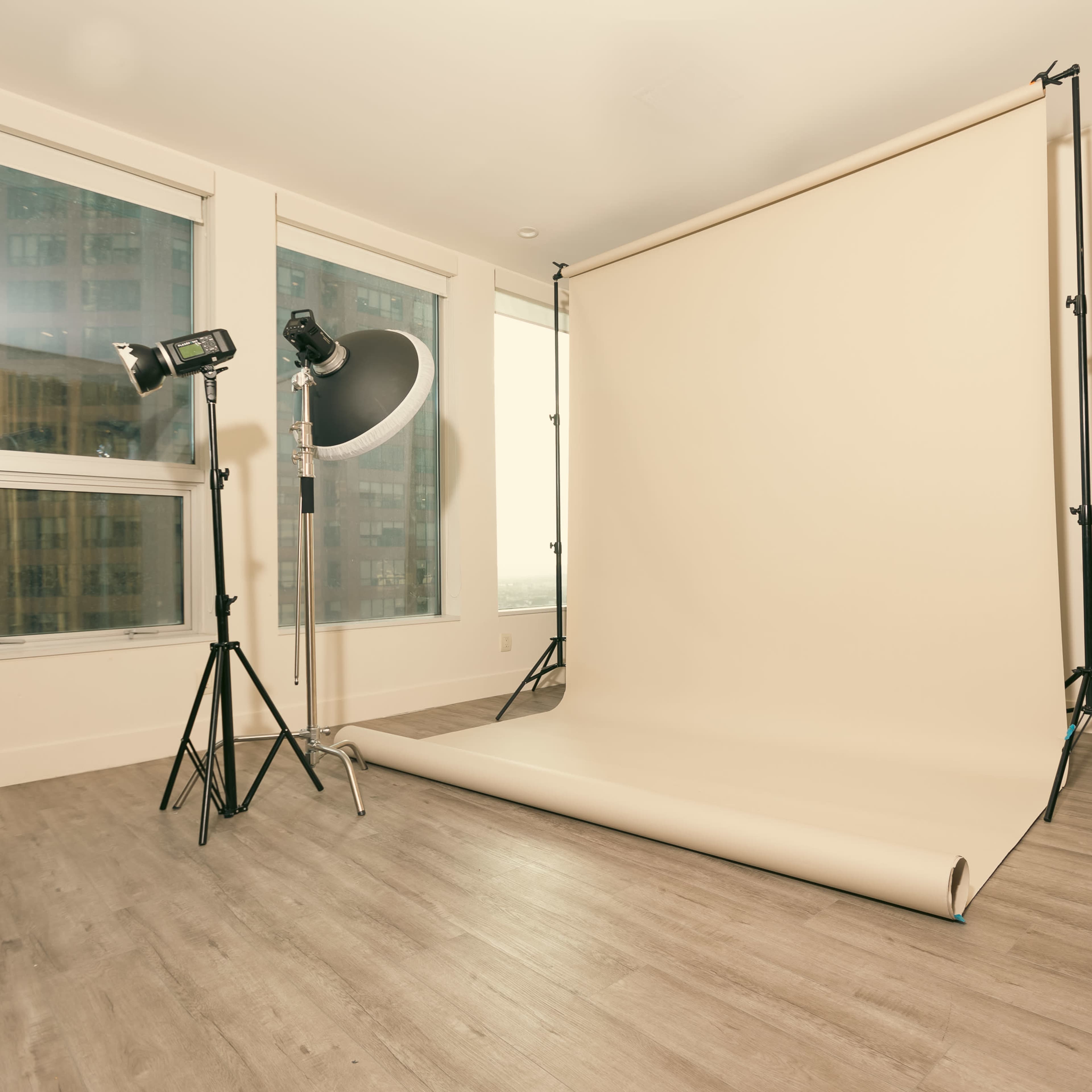 A photography studio with a beige backdrop set up on a smooth floor, accompanied by two studio lights and large windows in the background.