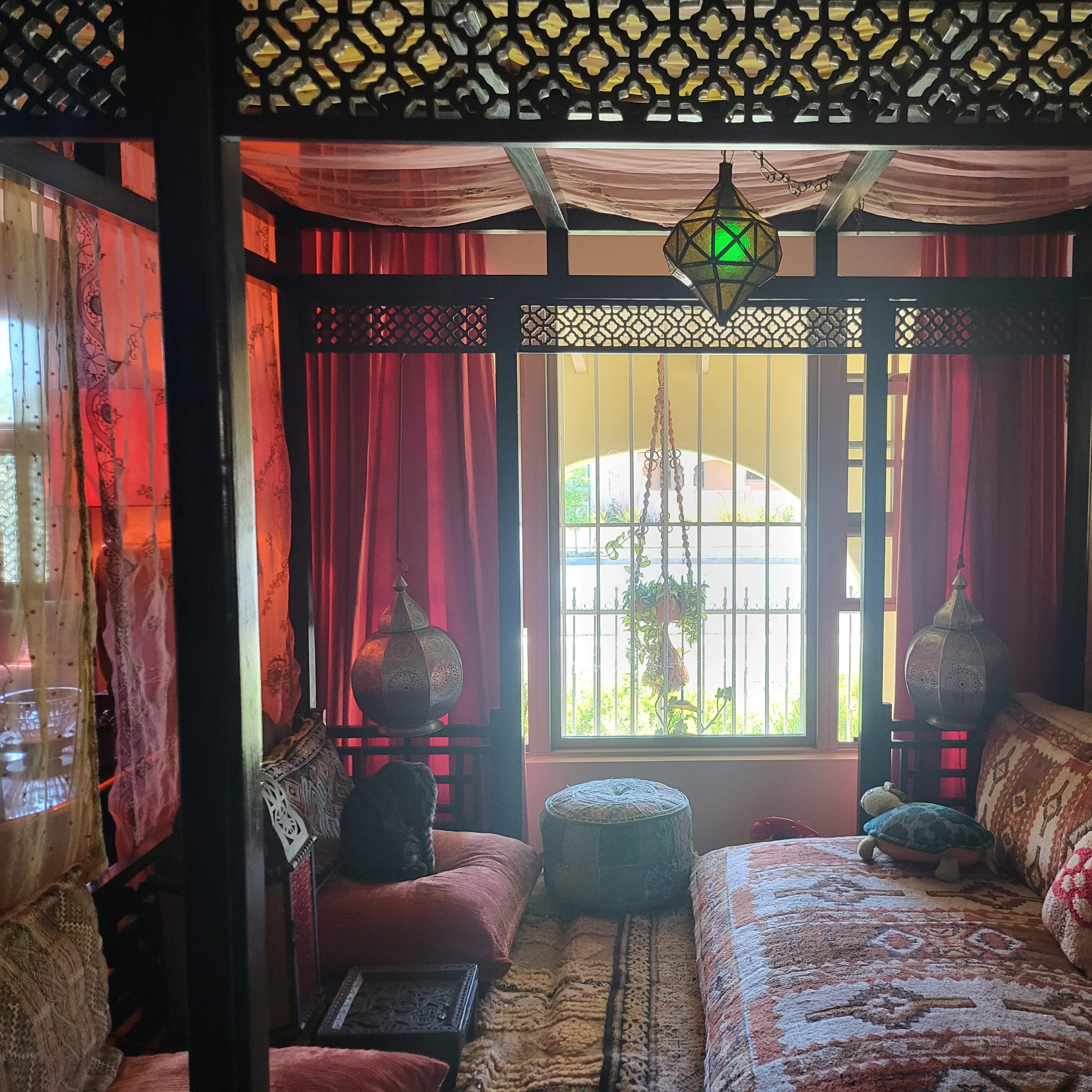 A cozy, elaborately decorated room featuring a four-poster bed with colorful textiles, lanterns, and patterned drapes, illuminated by natural light through a window.
