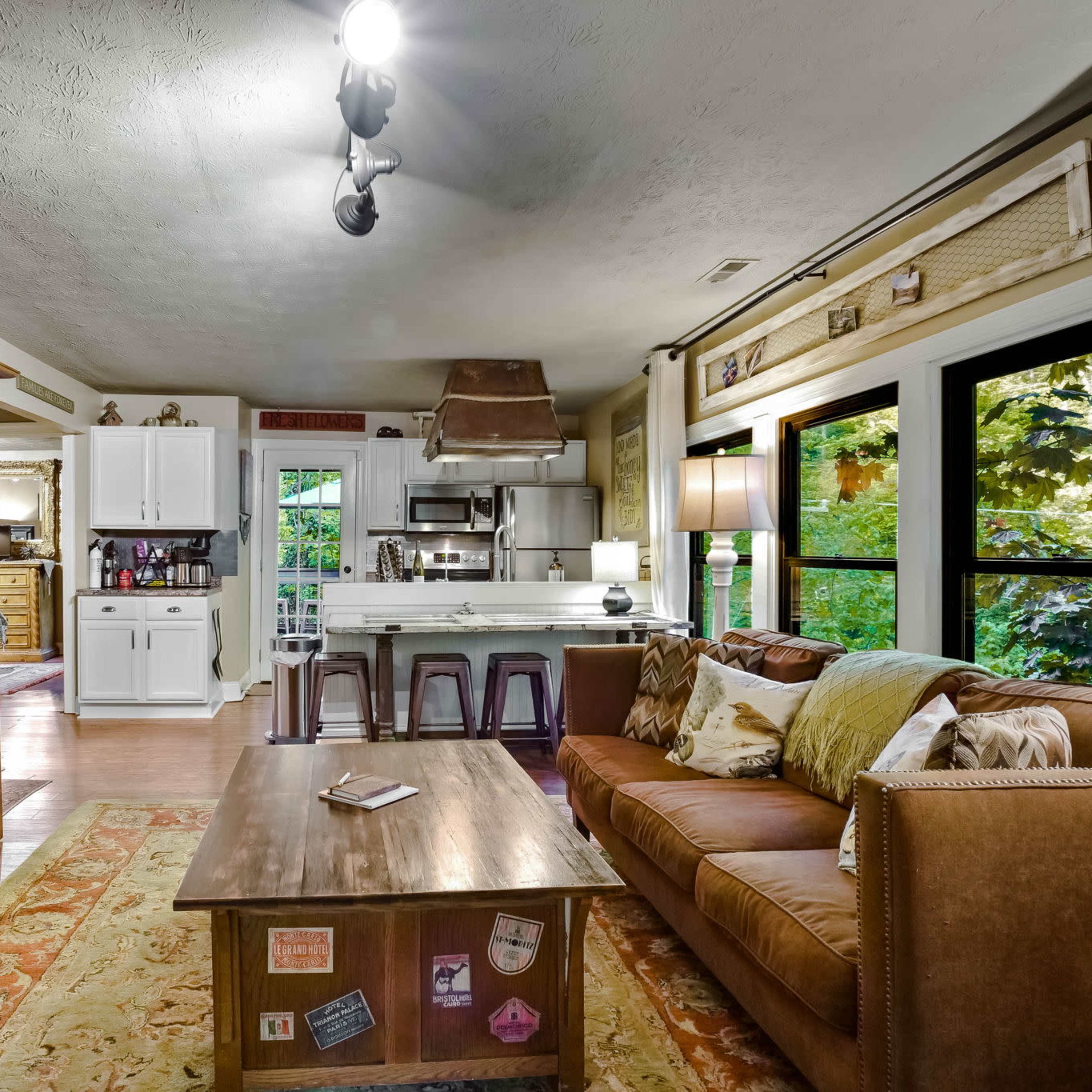 A living room features a brown sofa, a wooden coffee table, and a cabinet with a television, leading to an open kitchen in the background.