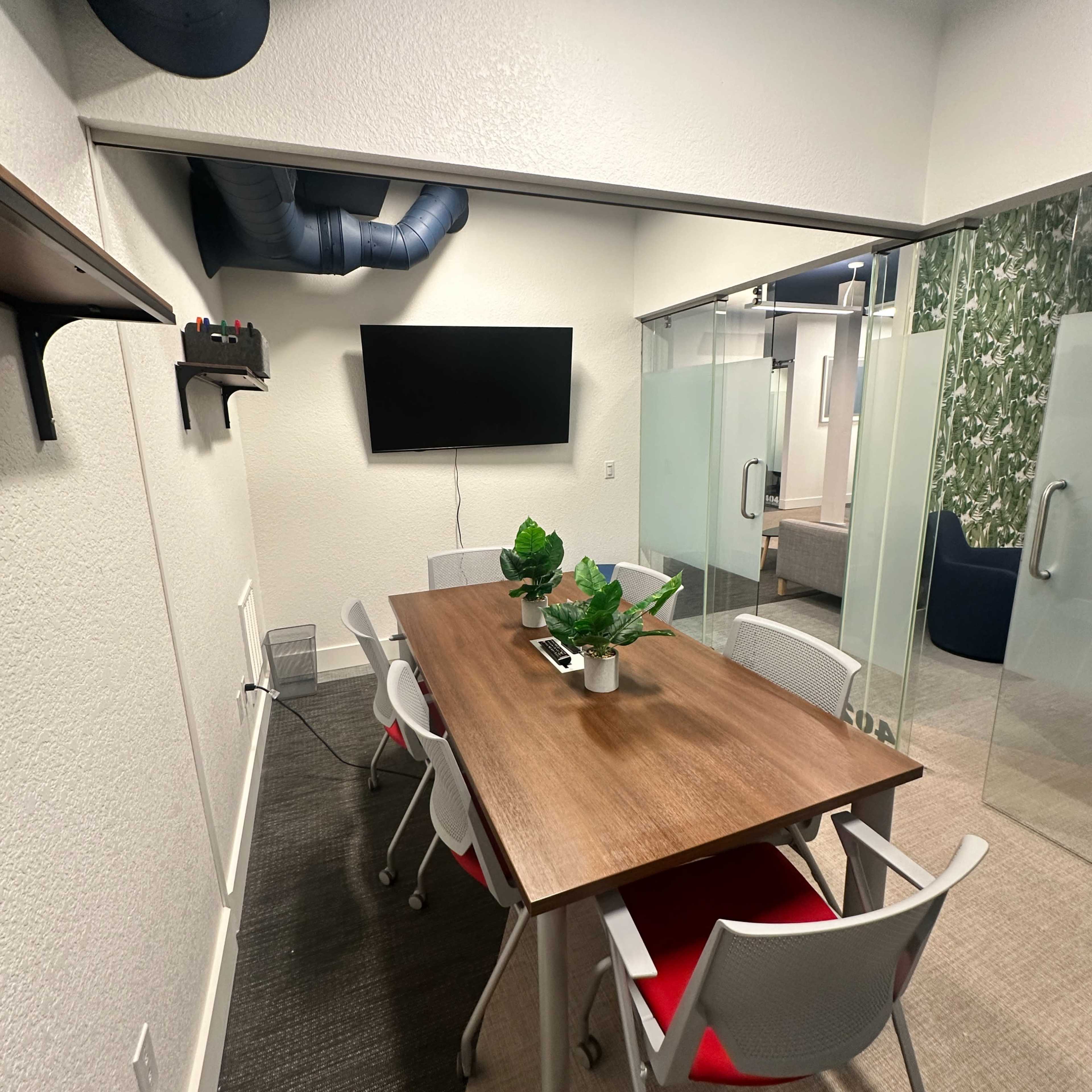 A modern conference room features a wooden table surrounded by four chairs, with a wall-mounted TV and decorative plants.