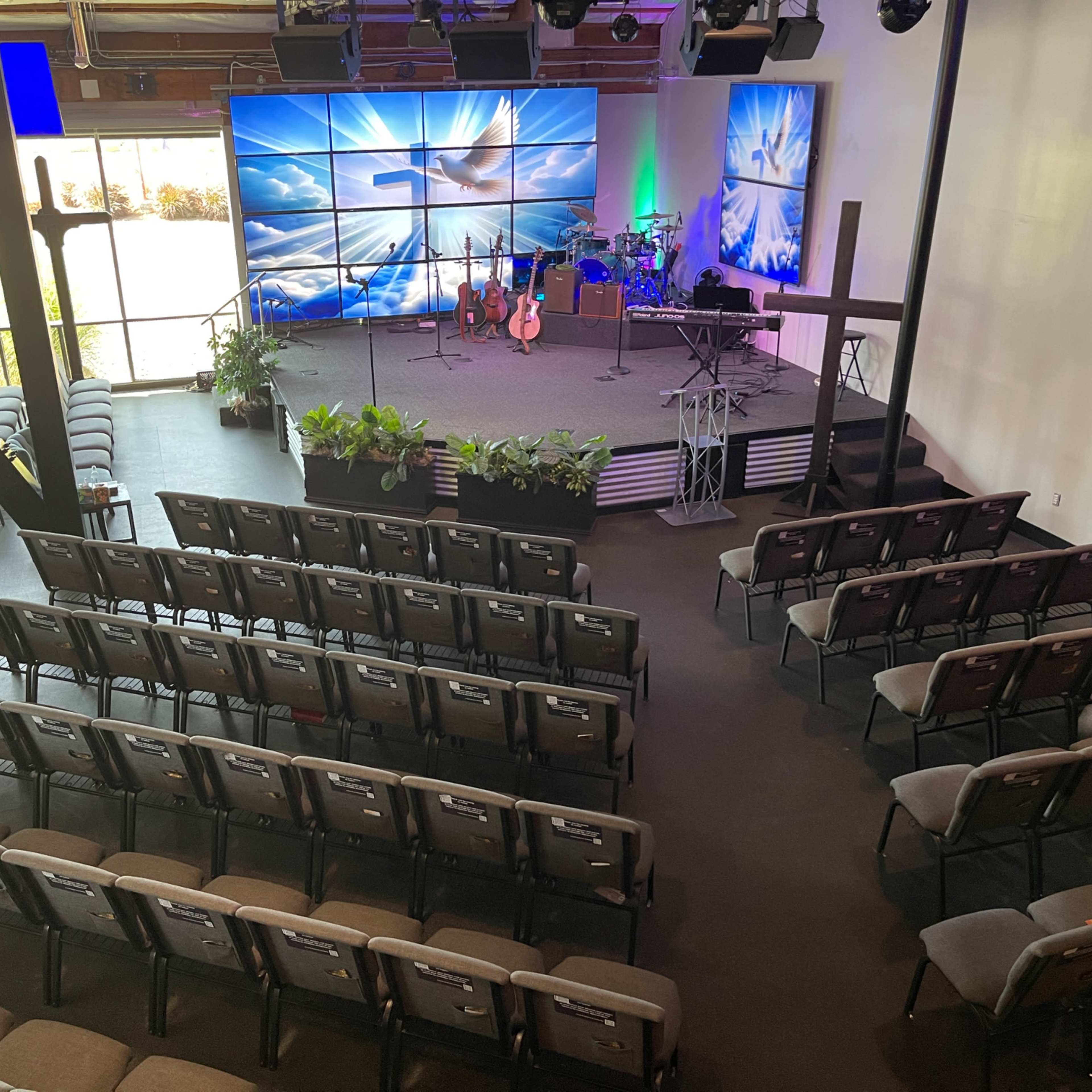A spacious auditorium features rows of seating facing a stage with musical instruments and a cross, illuminated by large screens displaying clouds and light.