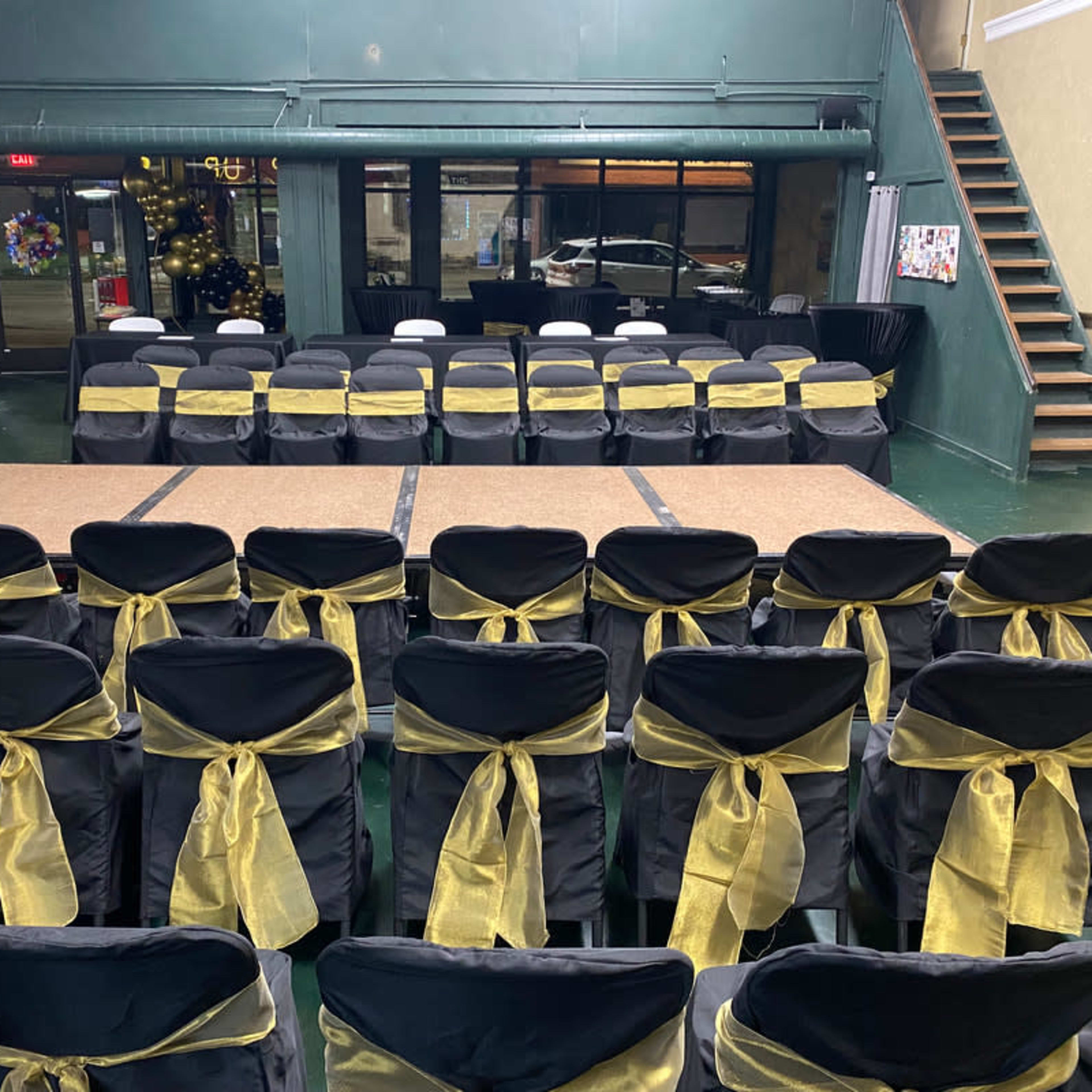 A spacious event venue set up with rows of black chairs adorned with gold sashes, facing a central stage area with a wooden platform.