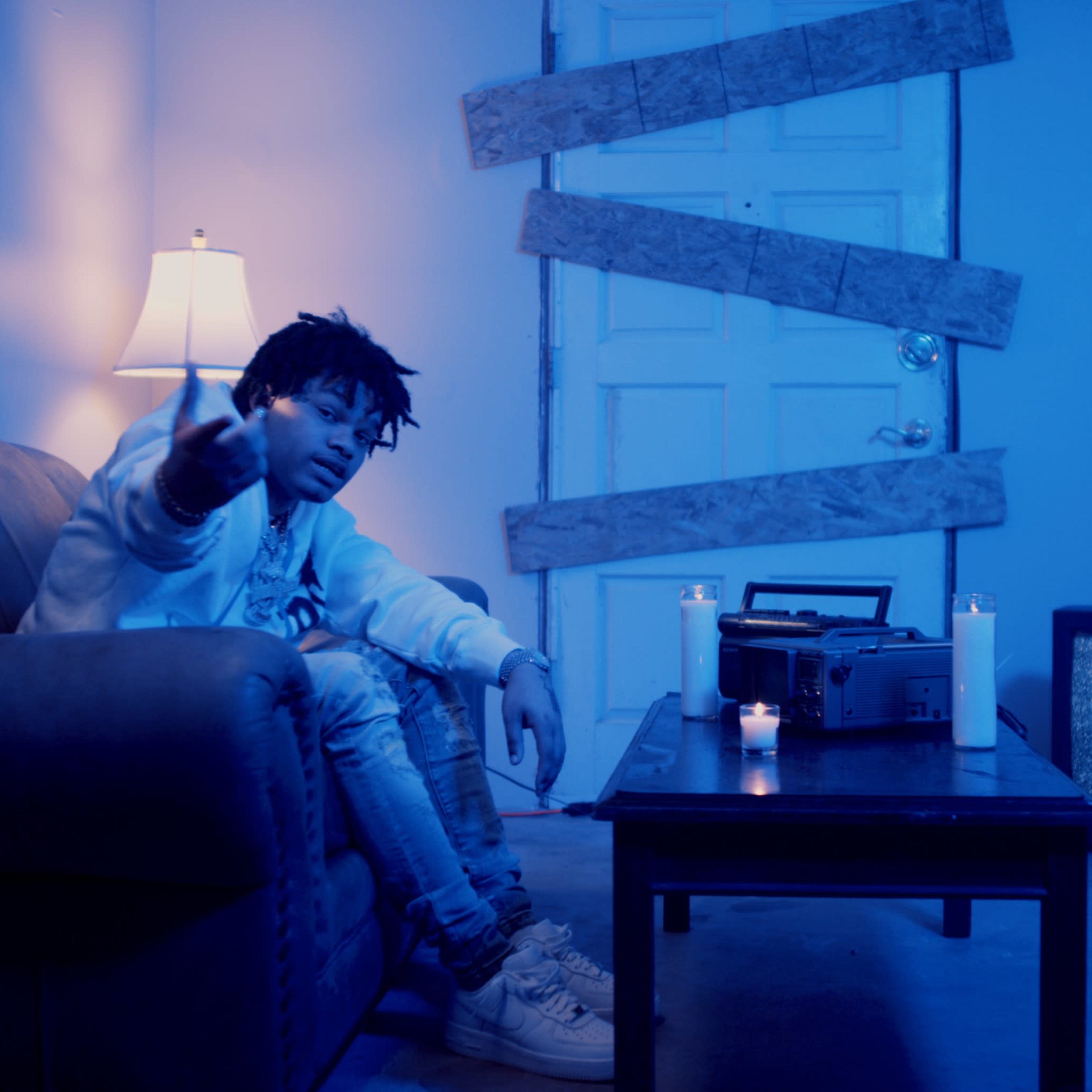 A person sits on a couch in a dimly lit room with a lamp, candles, and a recording device on a table, while wooden planks cover the door.