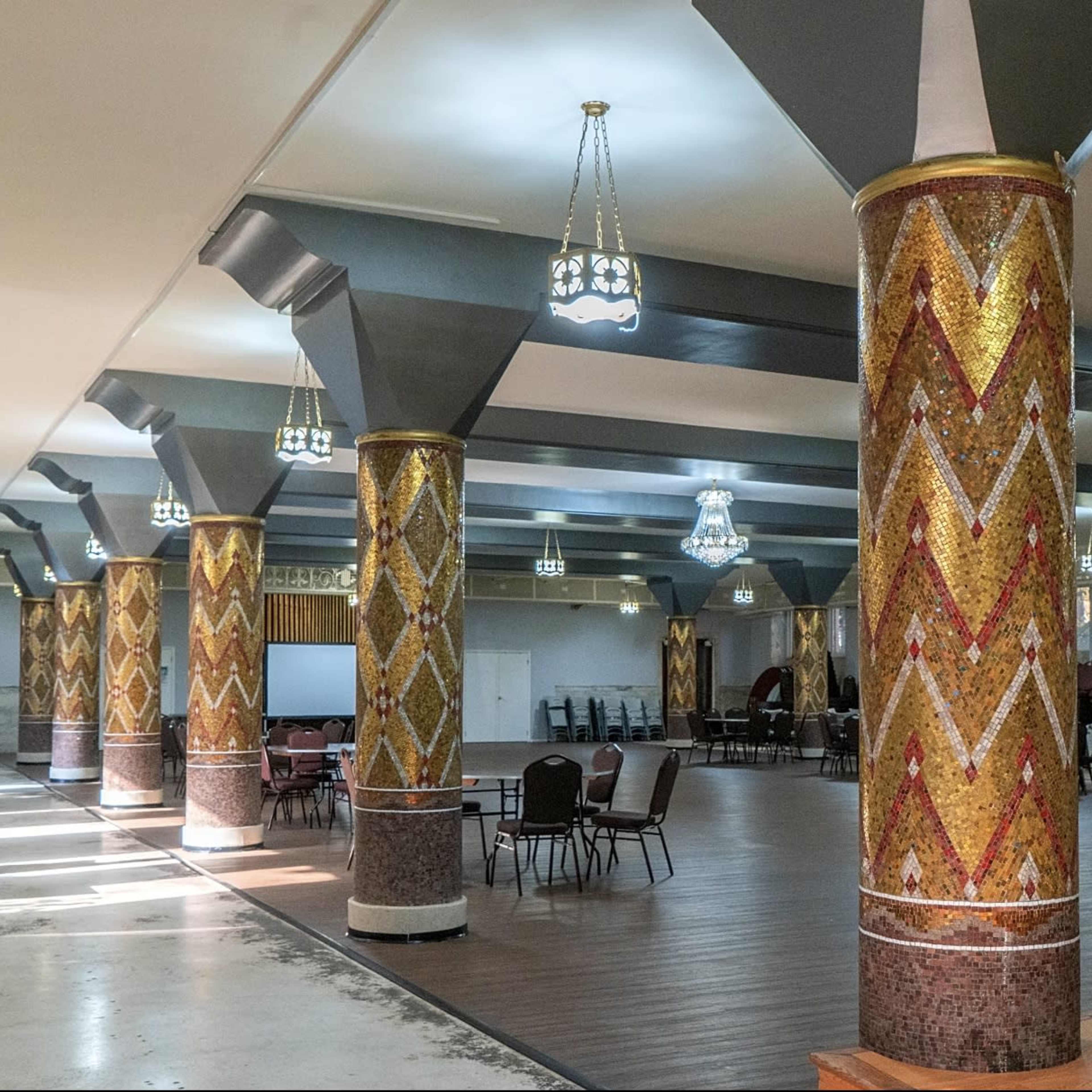 A spacious hall with decorative columns, chandeliers, and a seating arrangement, illuminated by natural light.