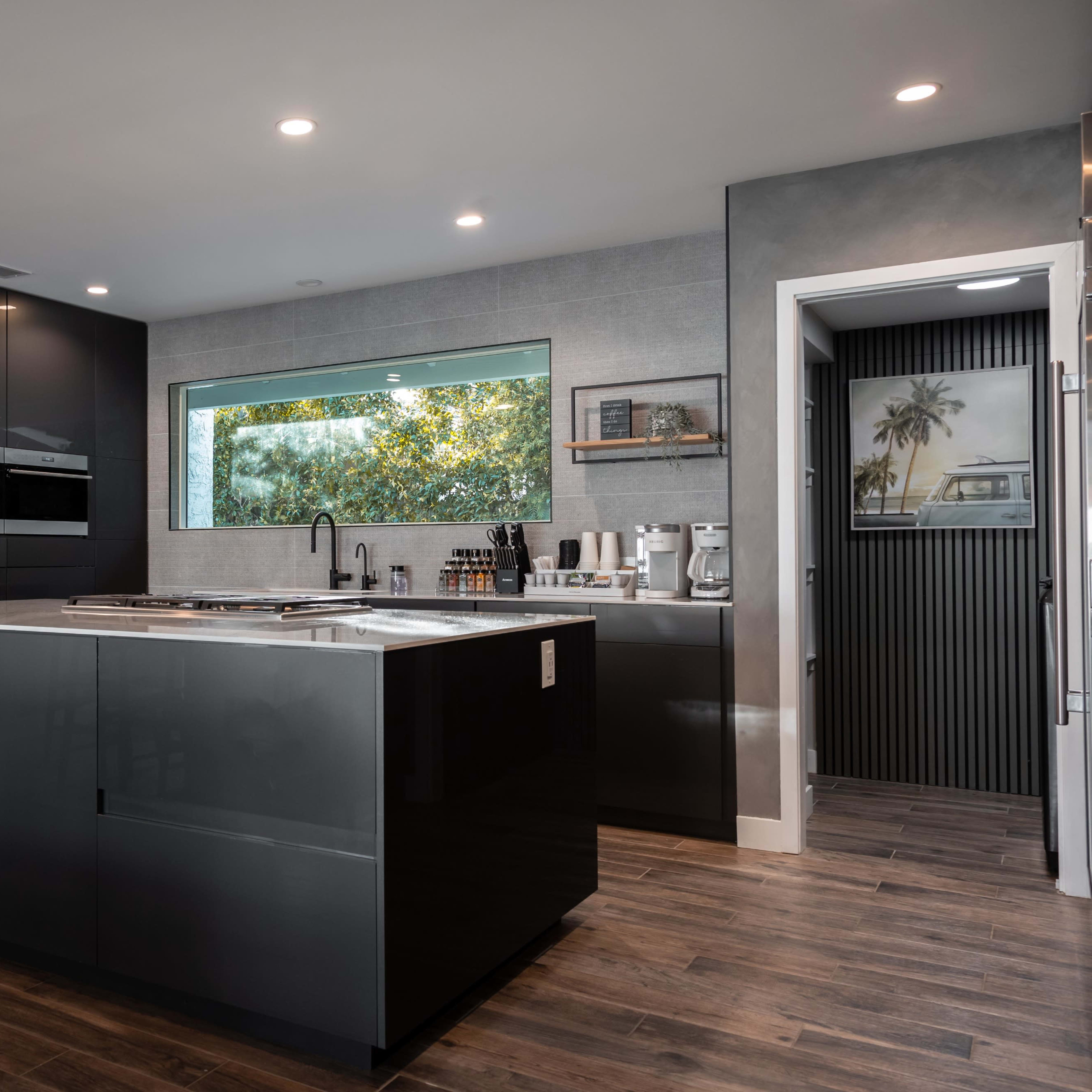 A modern kitchen features a large island with a sleek gray countertop, dark cabinetry, and a window overlooking greenery.
