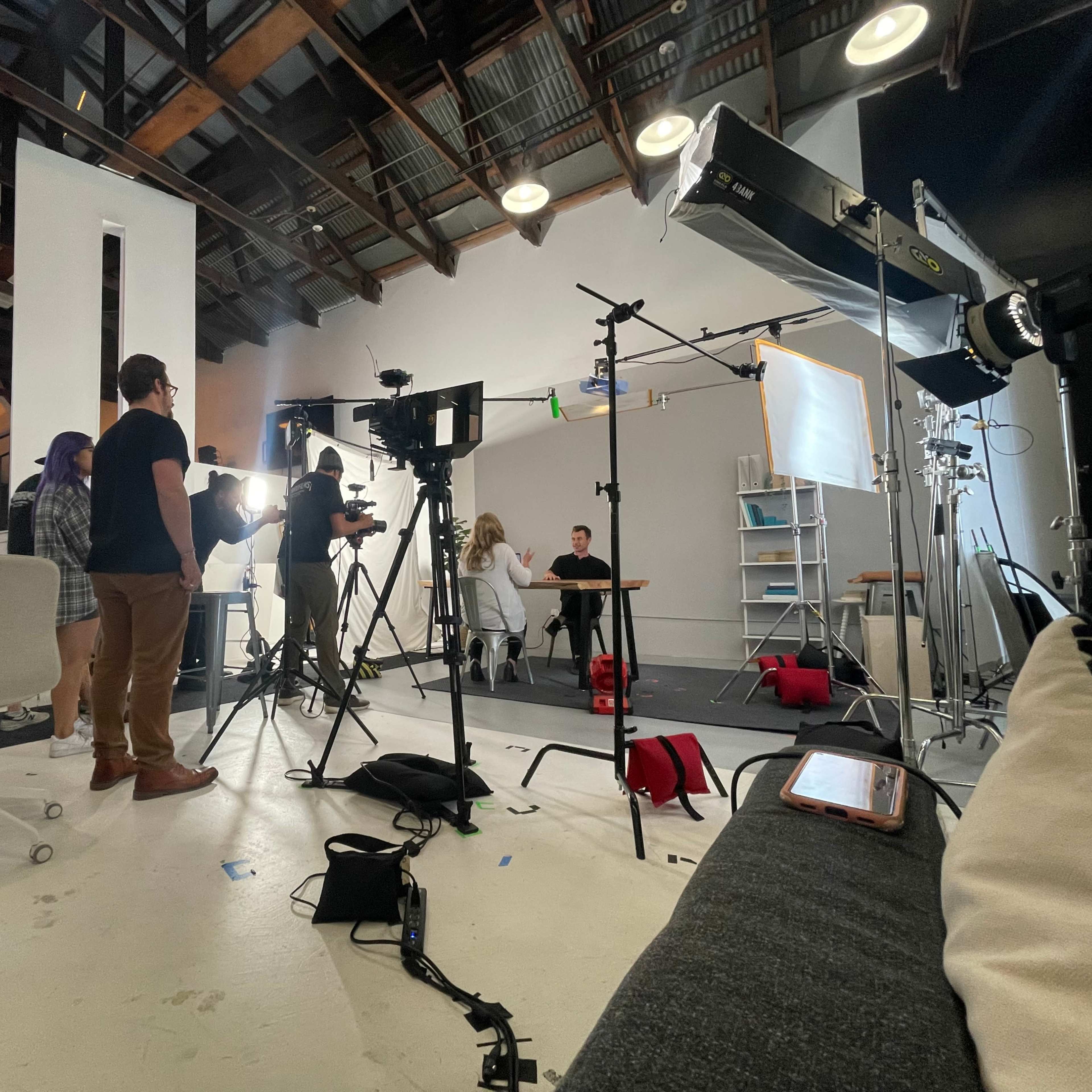 A film crew is set up in a studio, with equipment and lights arranged around two seated individuals at a table.