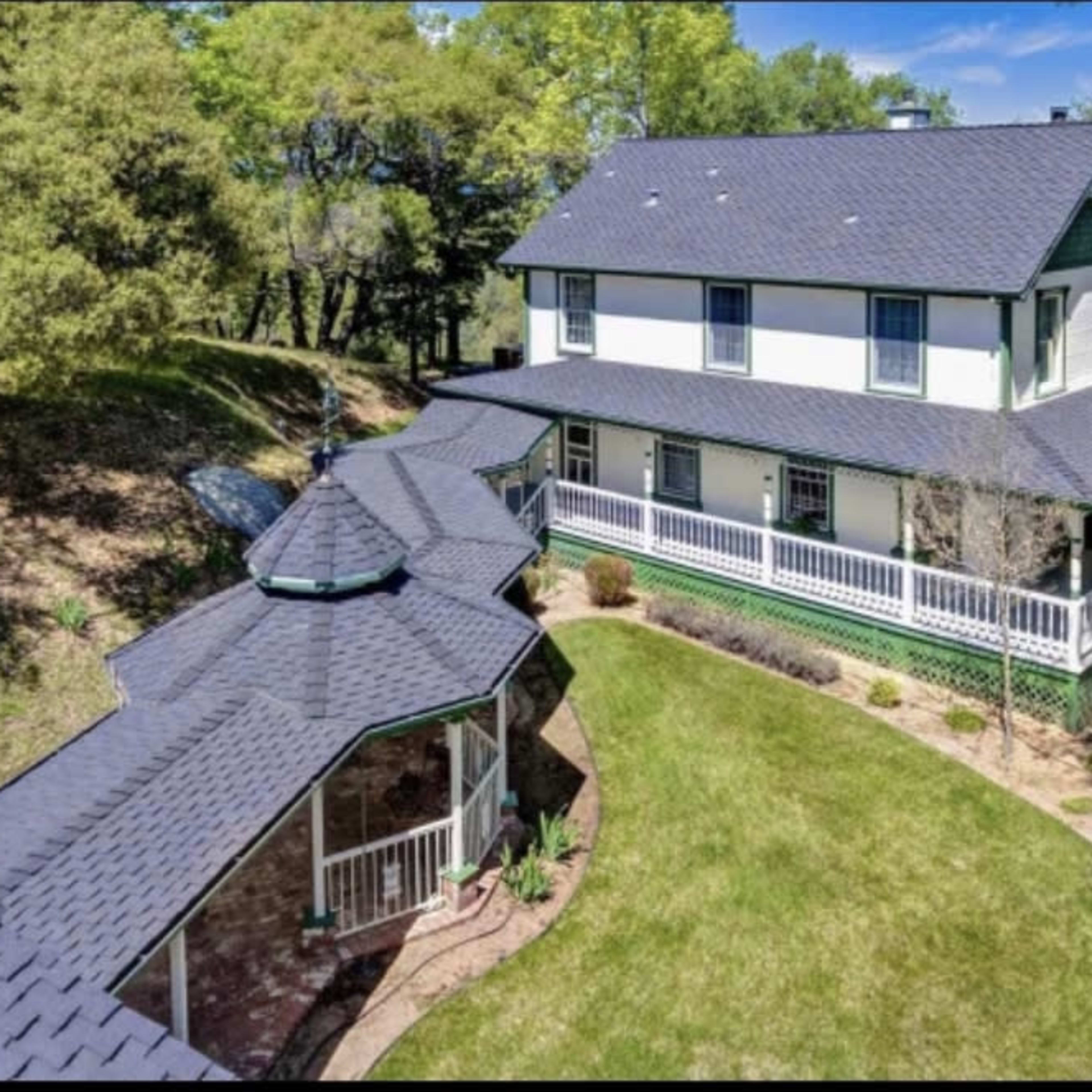 A two-story house with a large porch and a curved pathway is surrounded by trees and greenery.