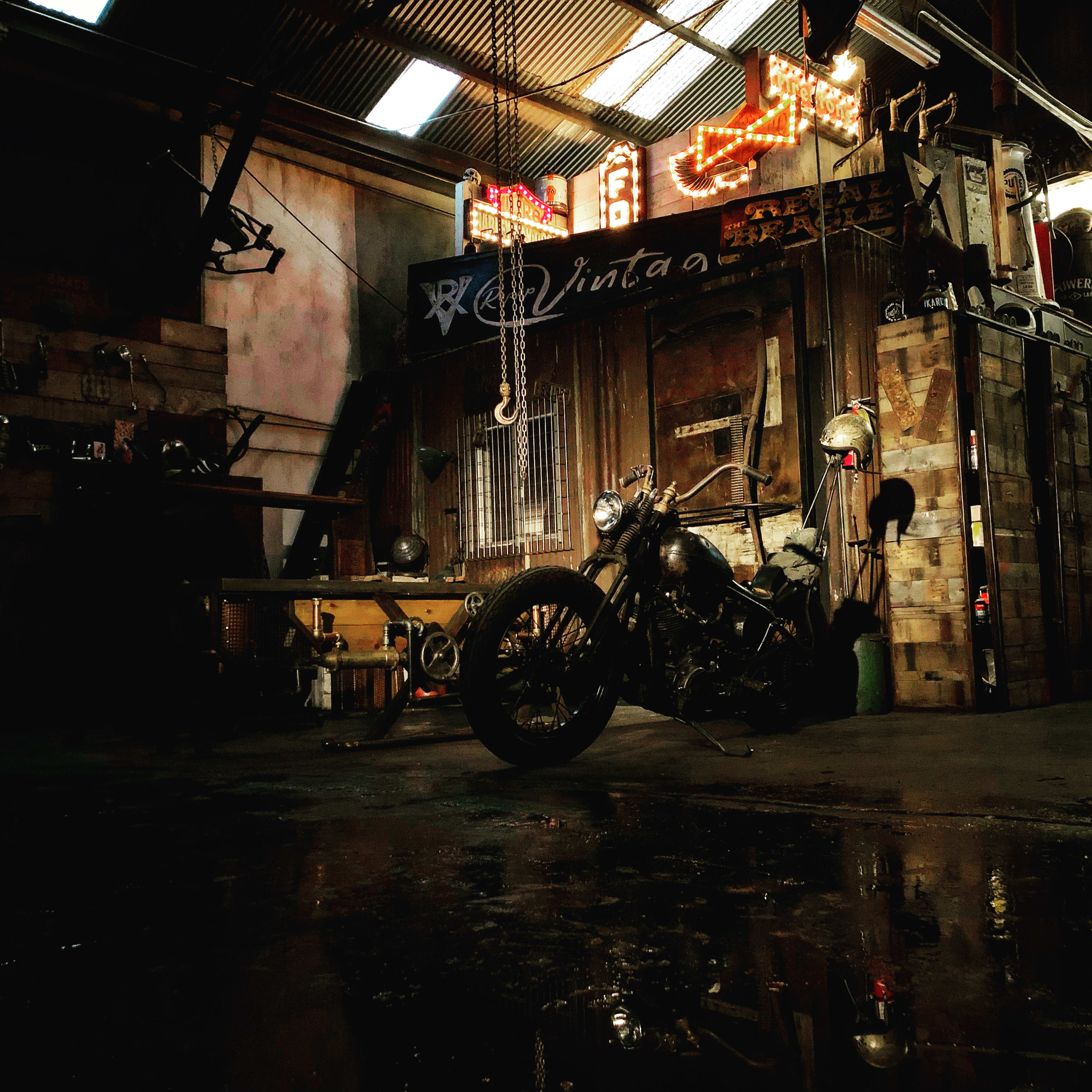 A vintage motorcycle stands in a dimly lit workshop with industrial decor and neon signs in the background.