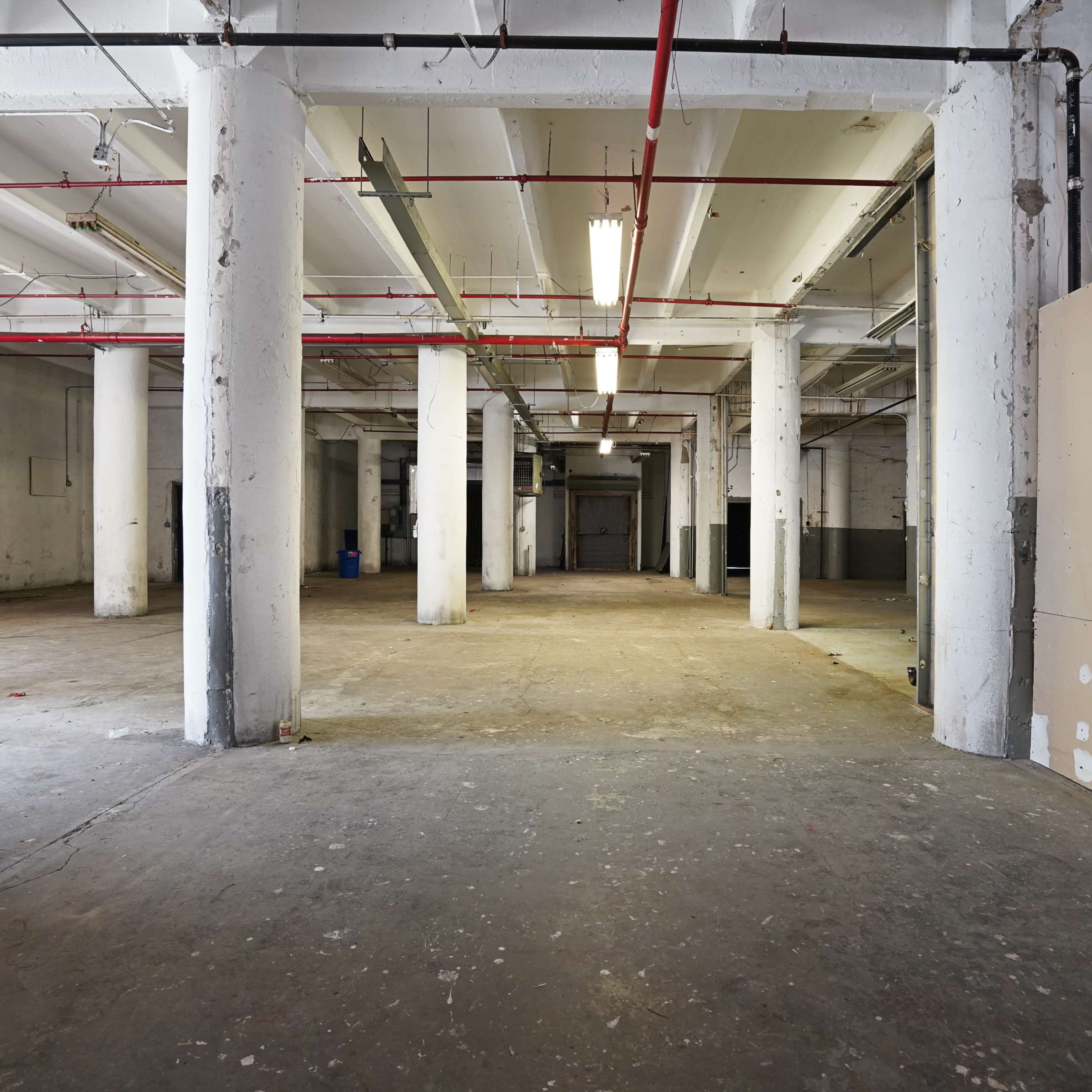 An empty industrial space with concrete floors, white columns, and exposed overhead piping.