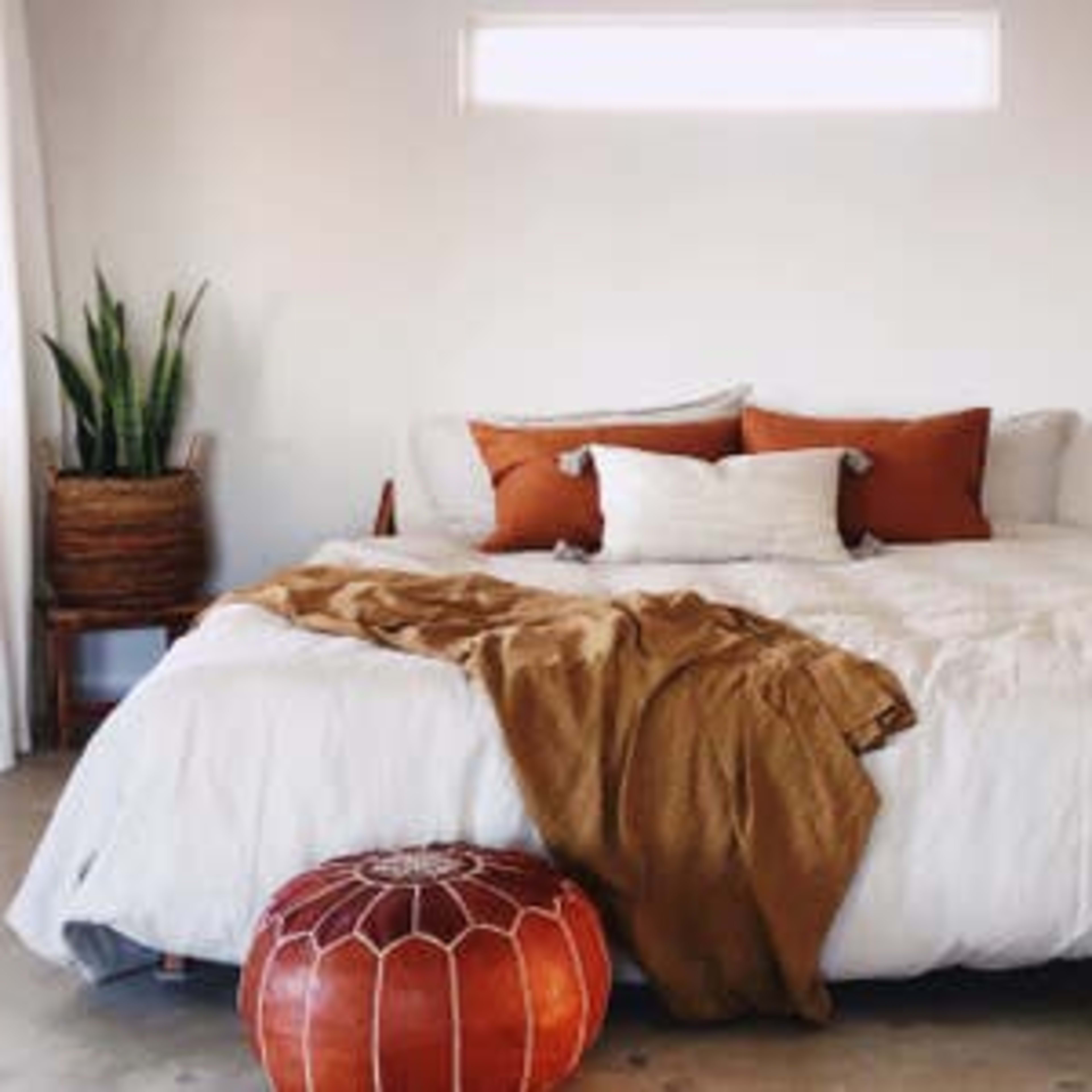 A neatly arranged bedroom features a large bed with white linens and several pillows, accompanied by a brown throw blanket and a decorative pouf.