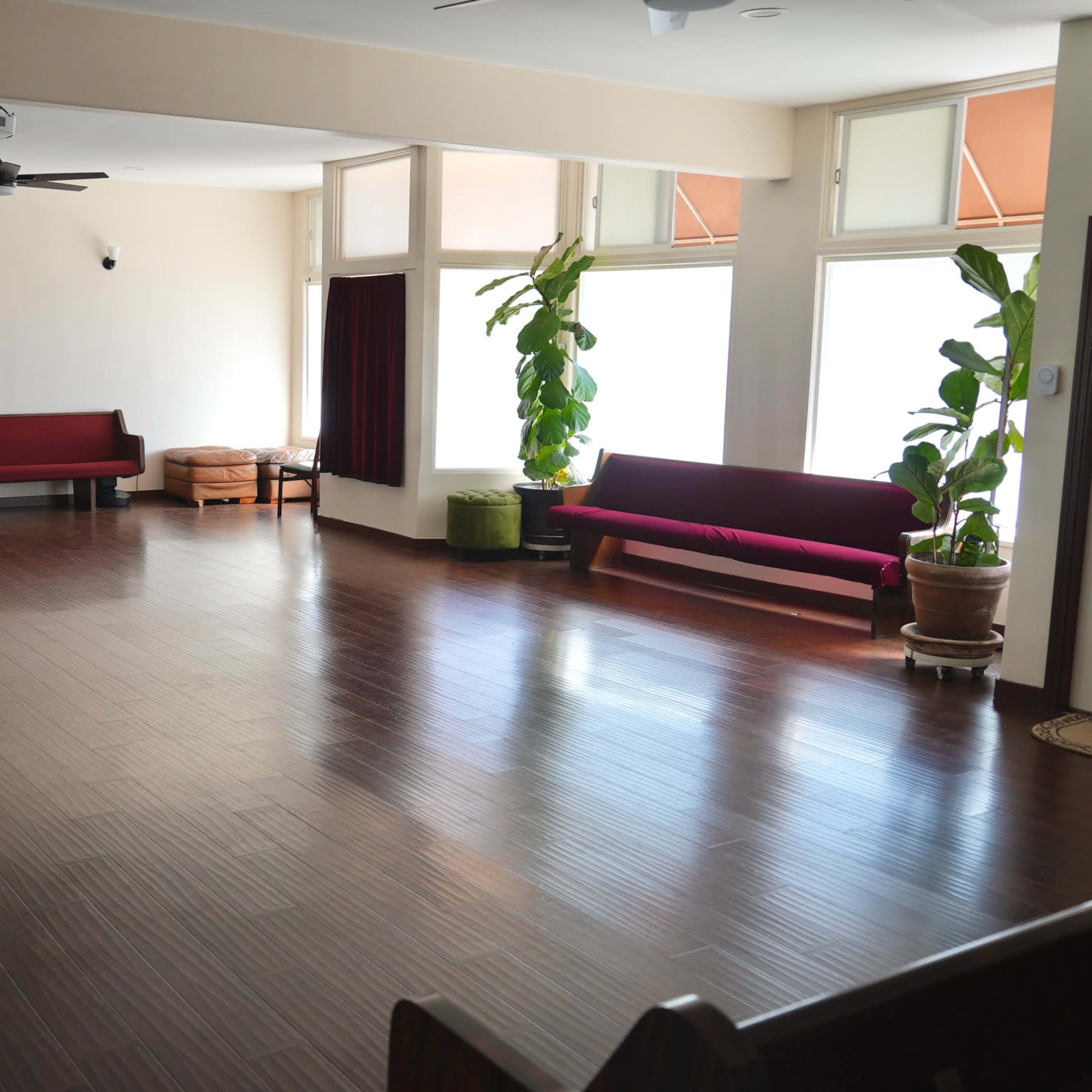 A spacious, empty room with wooden flooring, featuring two long benches along the walls and large windows allowing natural light to enter.