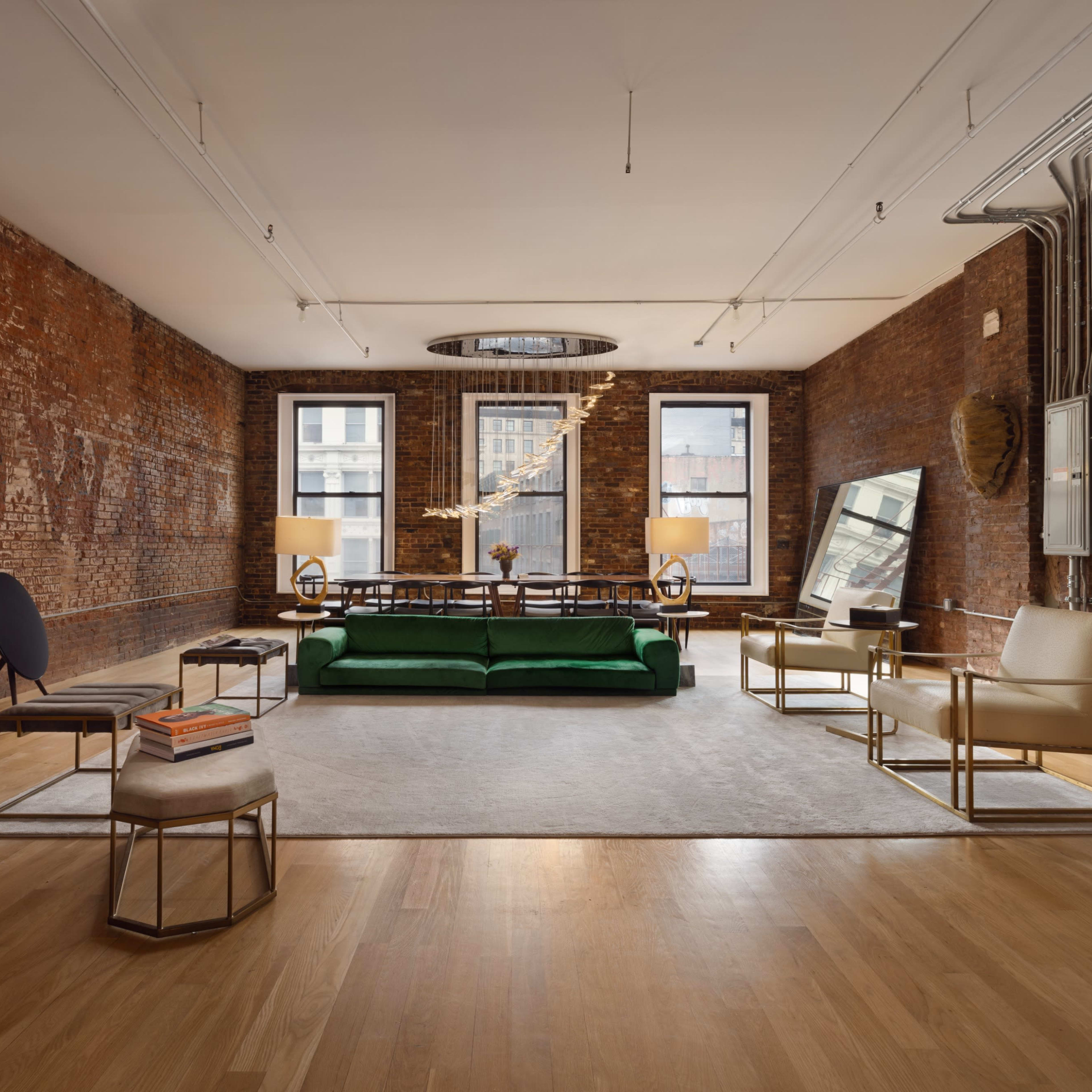 A modern living room with exposed brick walls, large windows, a green sofa, and minimalist furniture arranged on a light hardwood floor.
