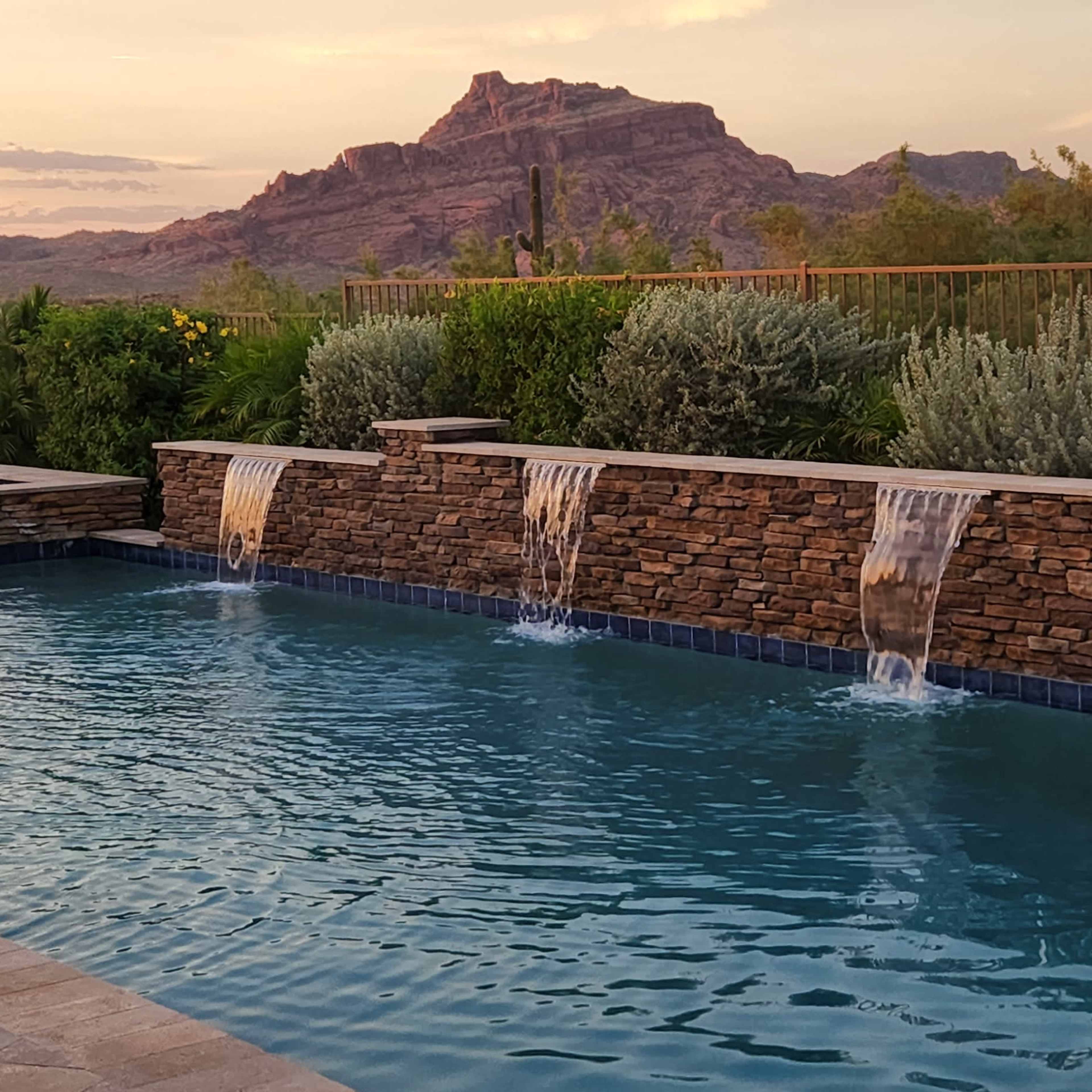A swimming pool with stone waterfalls is situated in a landscaped area, framed by mountains in the background during sunset.