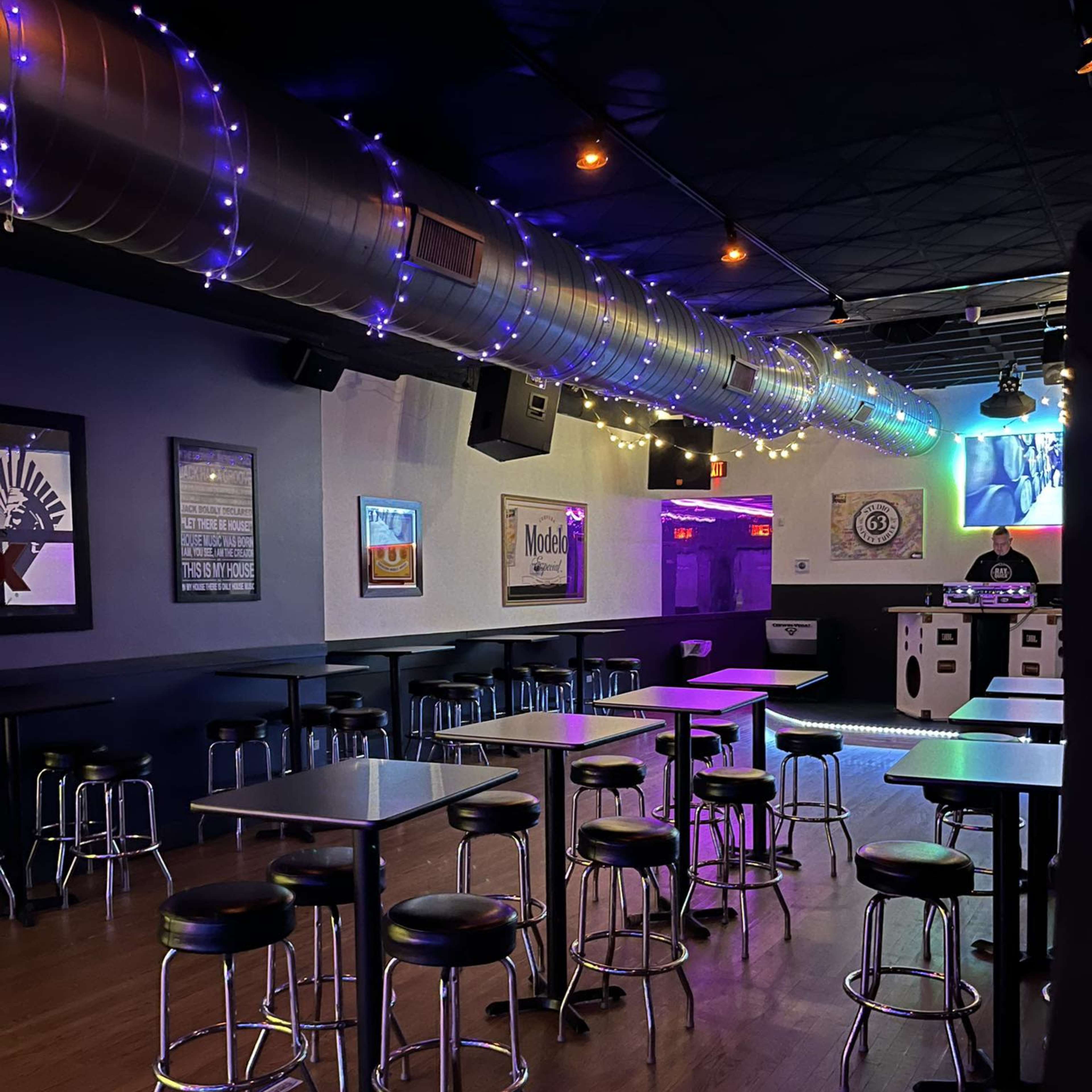 A dimly lit bar features rows of metal stools and tables, with colorful lights illuminating the space and a DJ booth in the background.