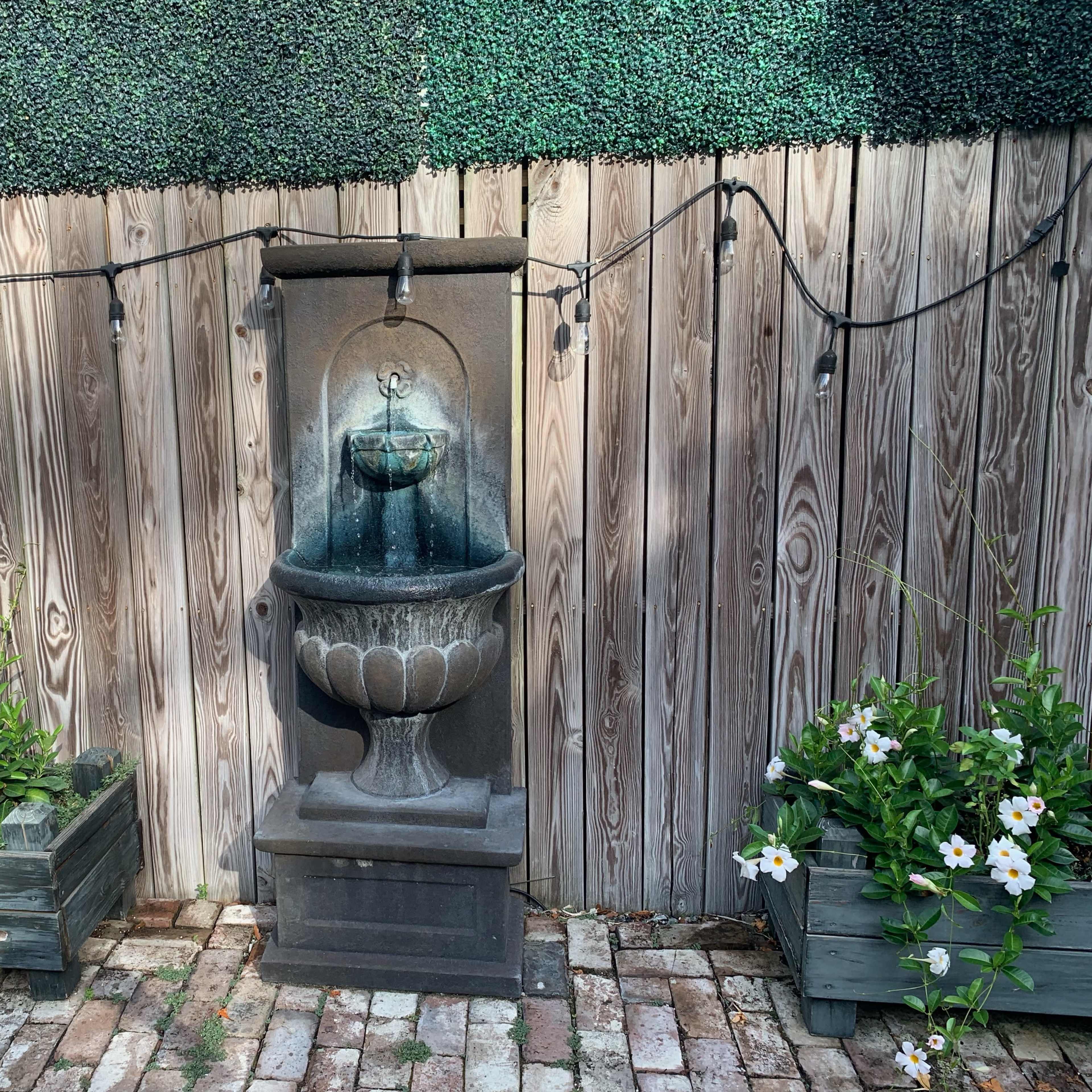 A stone fountain is positioned against a wooden fence, surrounded by two planter boxes filled with flowers and strung with decorative lights.