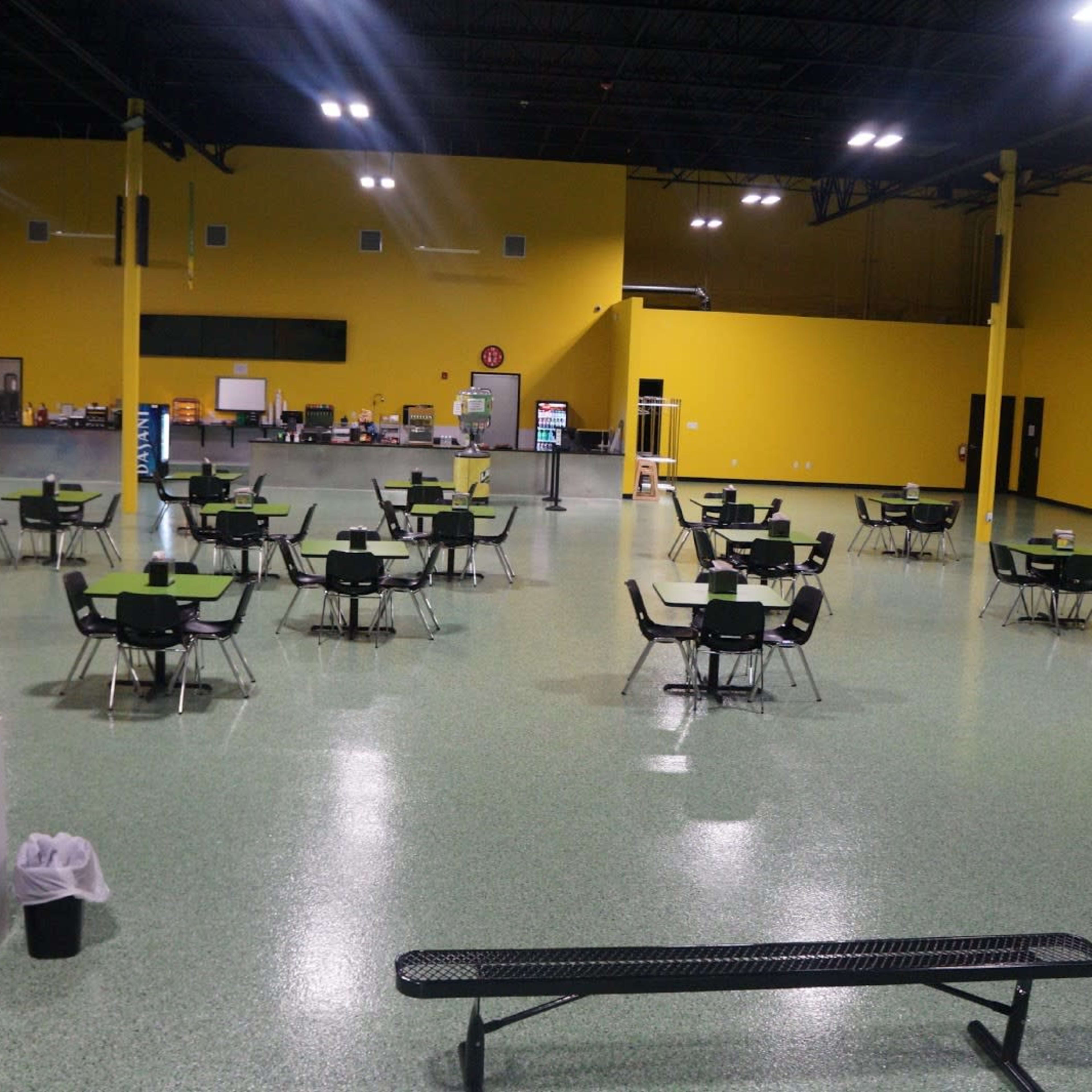 A large indoor space with multiple empty tables and chairs set up, surrounded by yellow walls and green flooring.