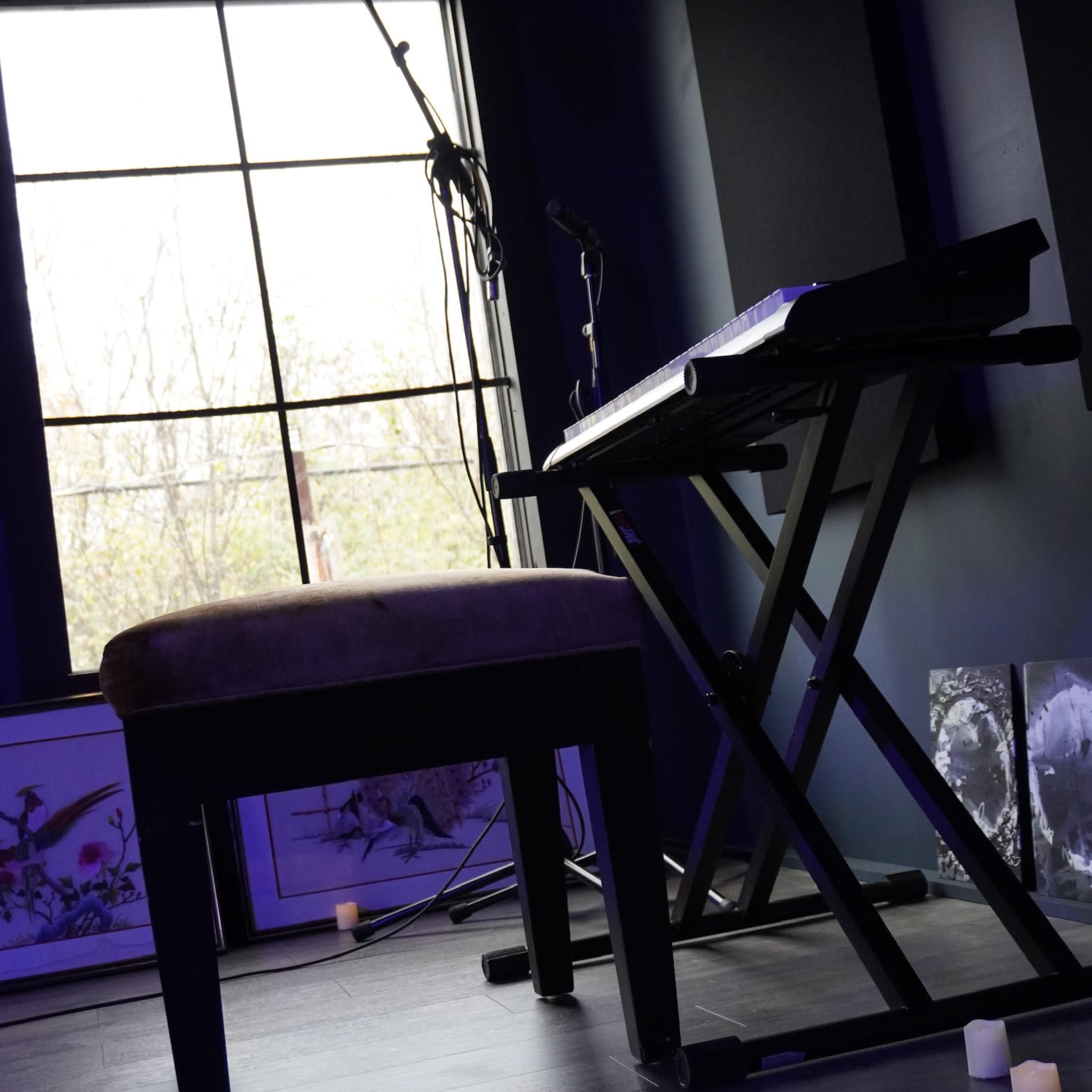 A microphone stands beside a keyboard and a stool in a dimly lit room with large windows.
