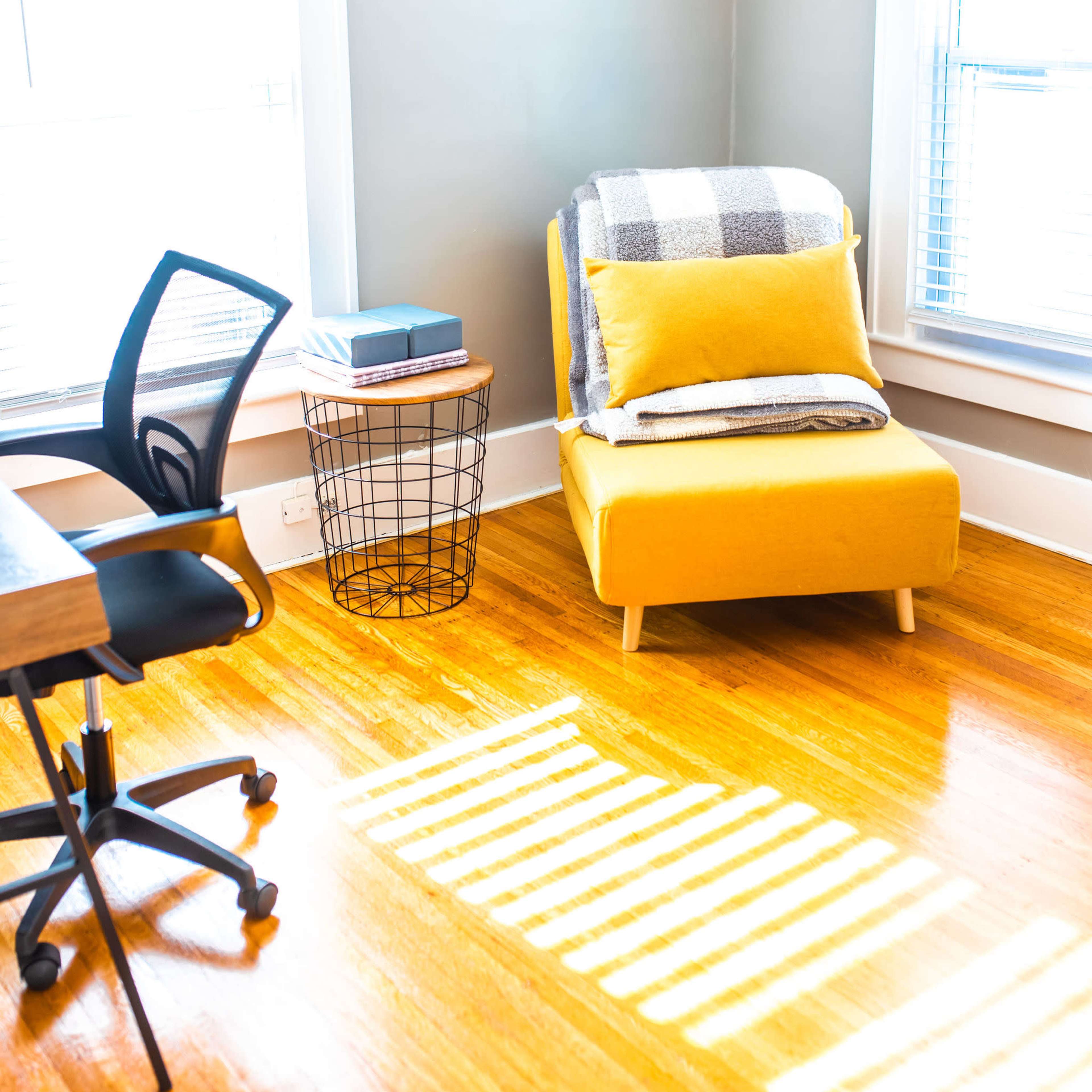 A bright corner of a room featuring a yellow chair with a blanket, a side table with a book, and a black office chair positioned near a wooden desk.
