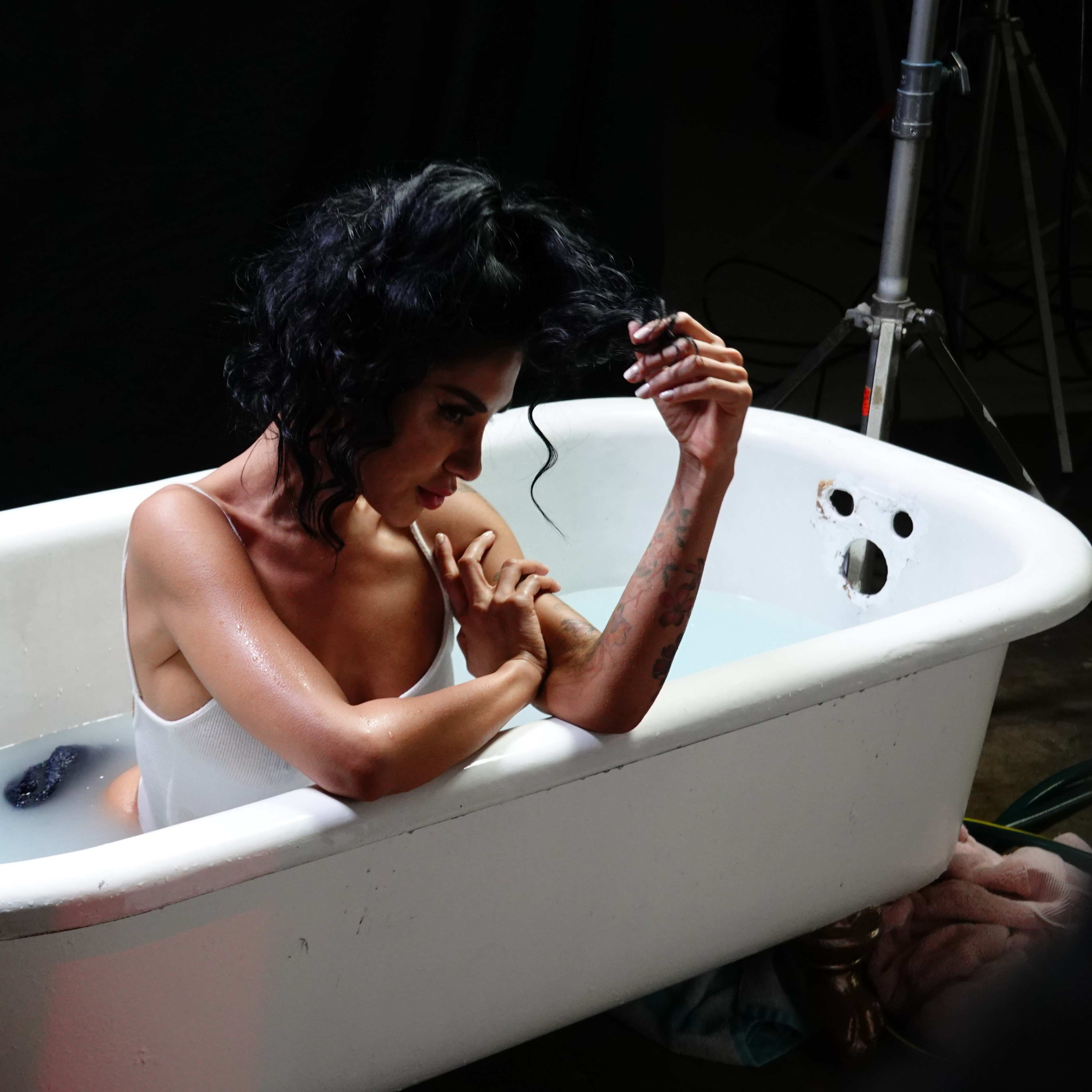 A woman with dark hair sits in a white bathtub filled with water, holding her arm while looking contemplatively towards the side.