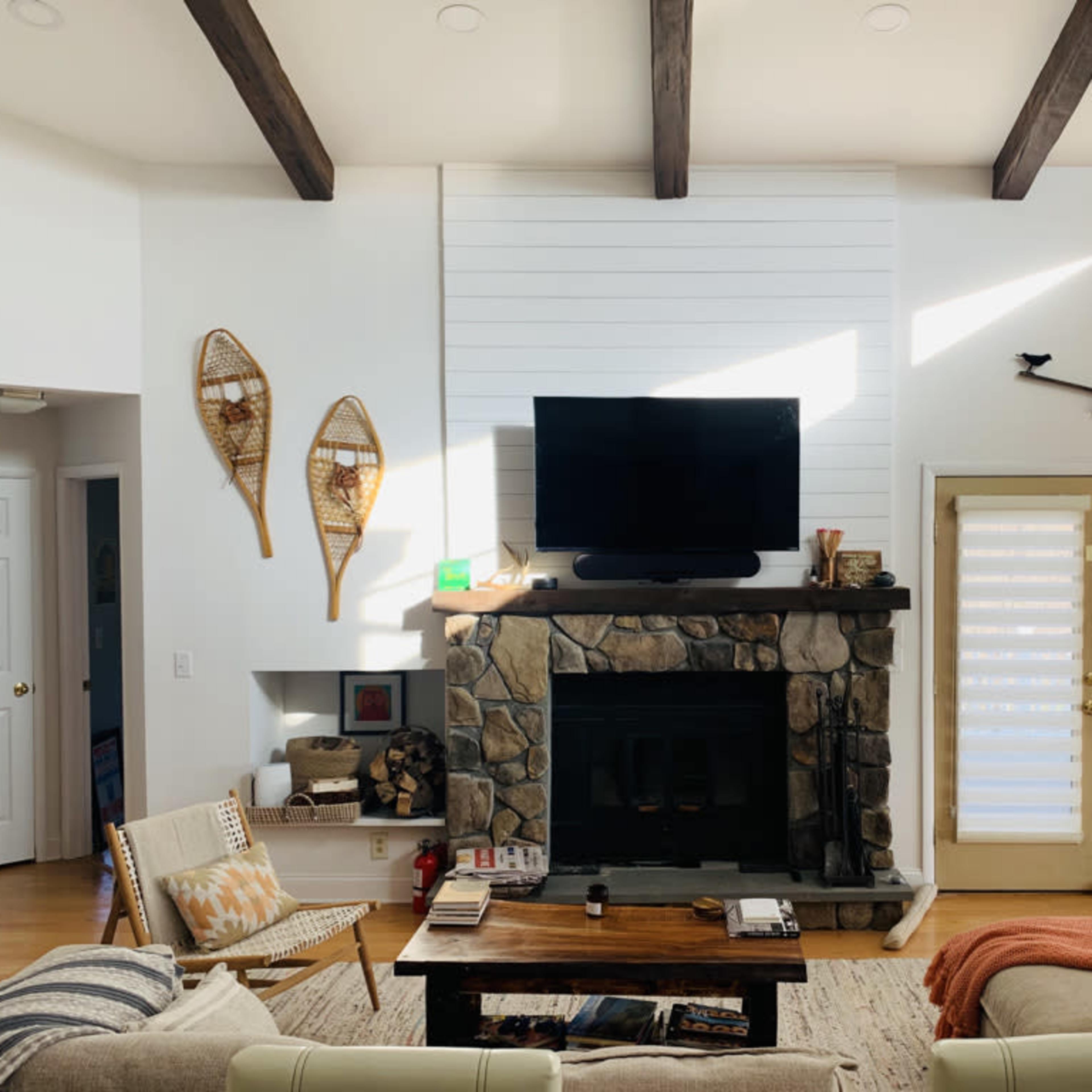 A cozy living room with a stone fireplace, a mounted television above it, and large glass doors leading outside, complemented by wooden beams on the ceiling.