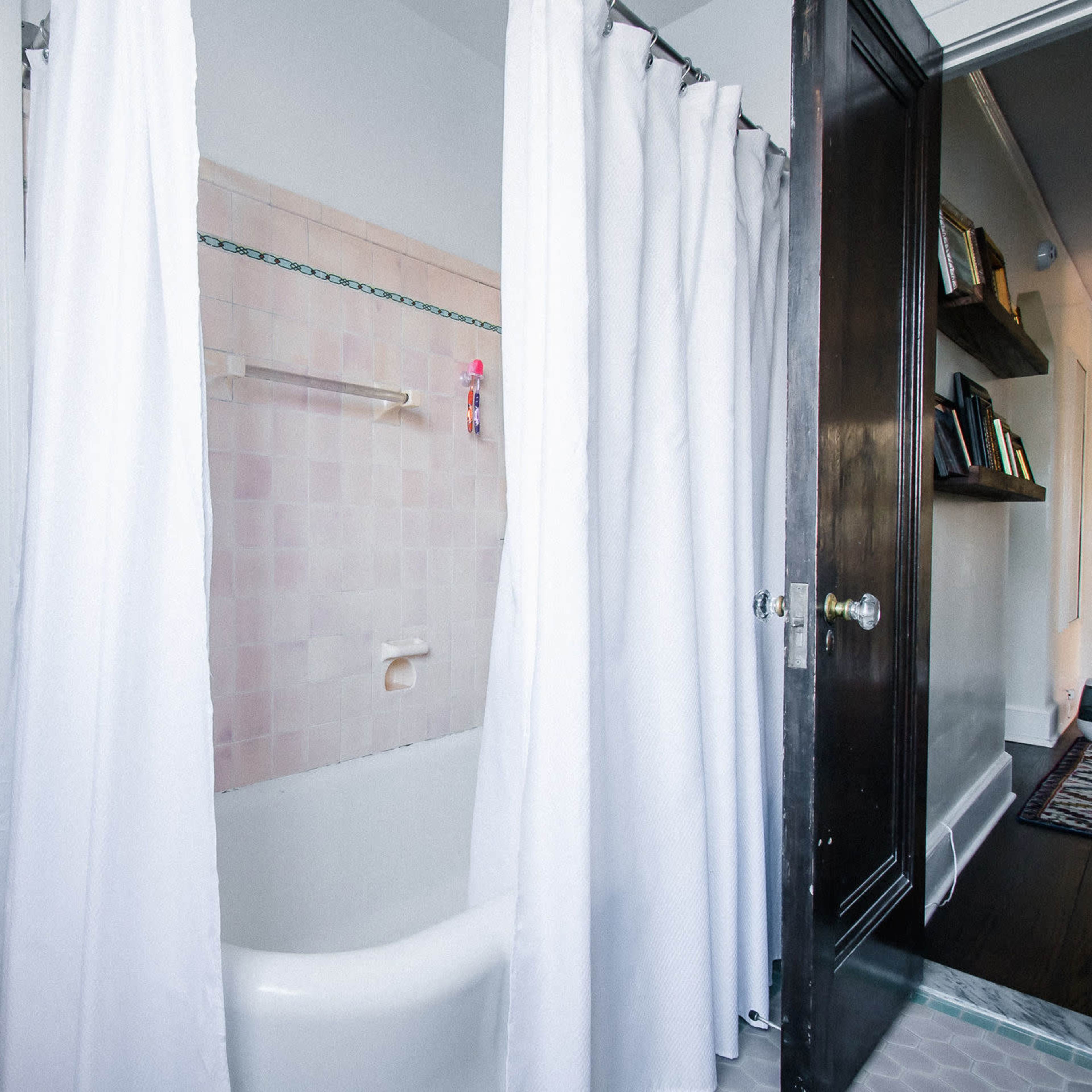 A partially visible bathroom with a bathtub surrounded by white curtains, leading to a well-lit area with a chair and a bookshelf in the background.