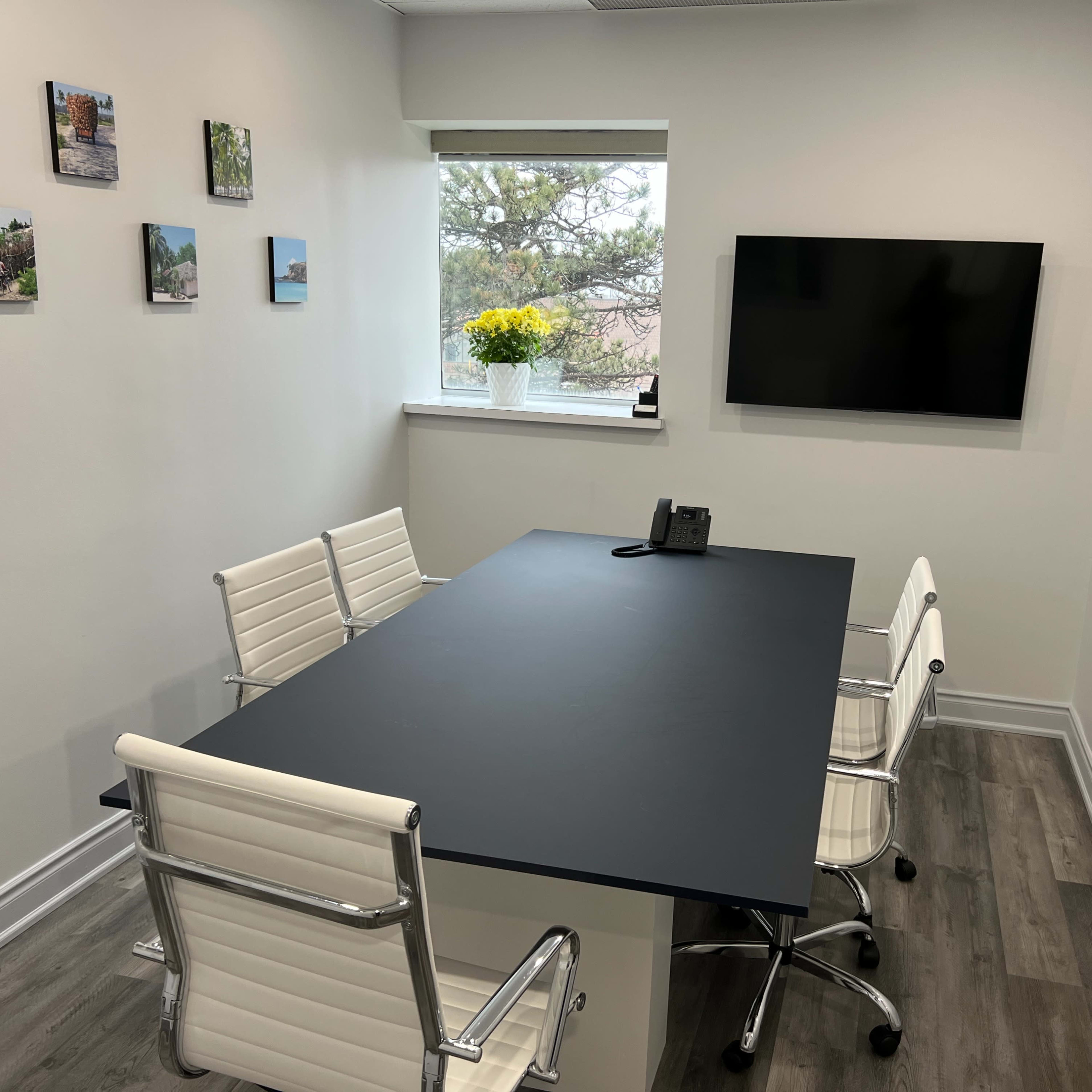 A conference room features a black table, four white chairs, a phone, and a television mounted on the wall, with framed photos arranged around the room and a window with flowers.