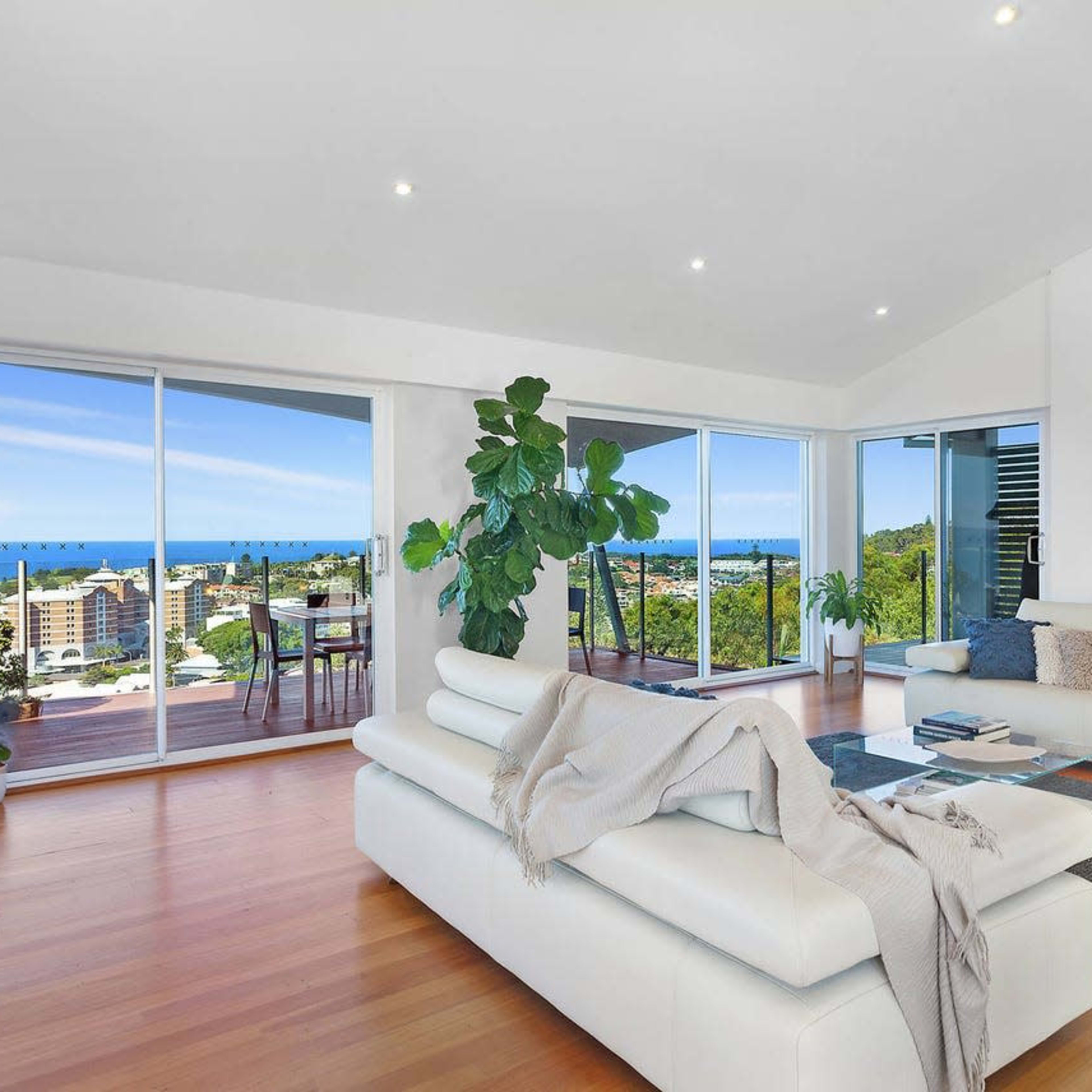 A modern living room with large windows offering a view of the ocean and a balcony area, featuring a white sofa, a large plant, and wooden flooring.