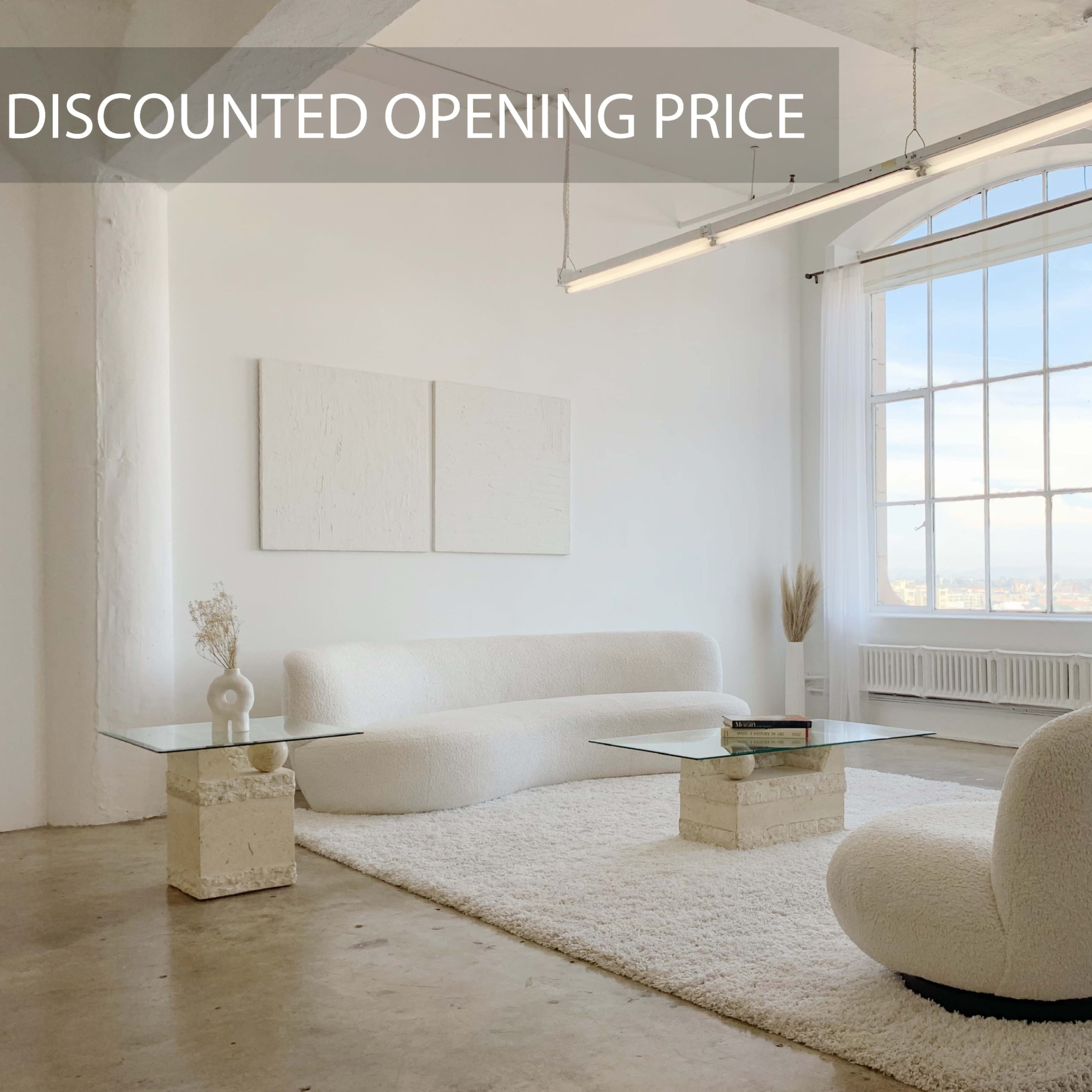 A spacious, minimalist living room featuring a white, plush sofa, two rounded chairs, and a glass coffee table, all set against a backdrop of large windows.