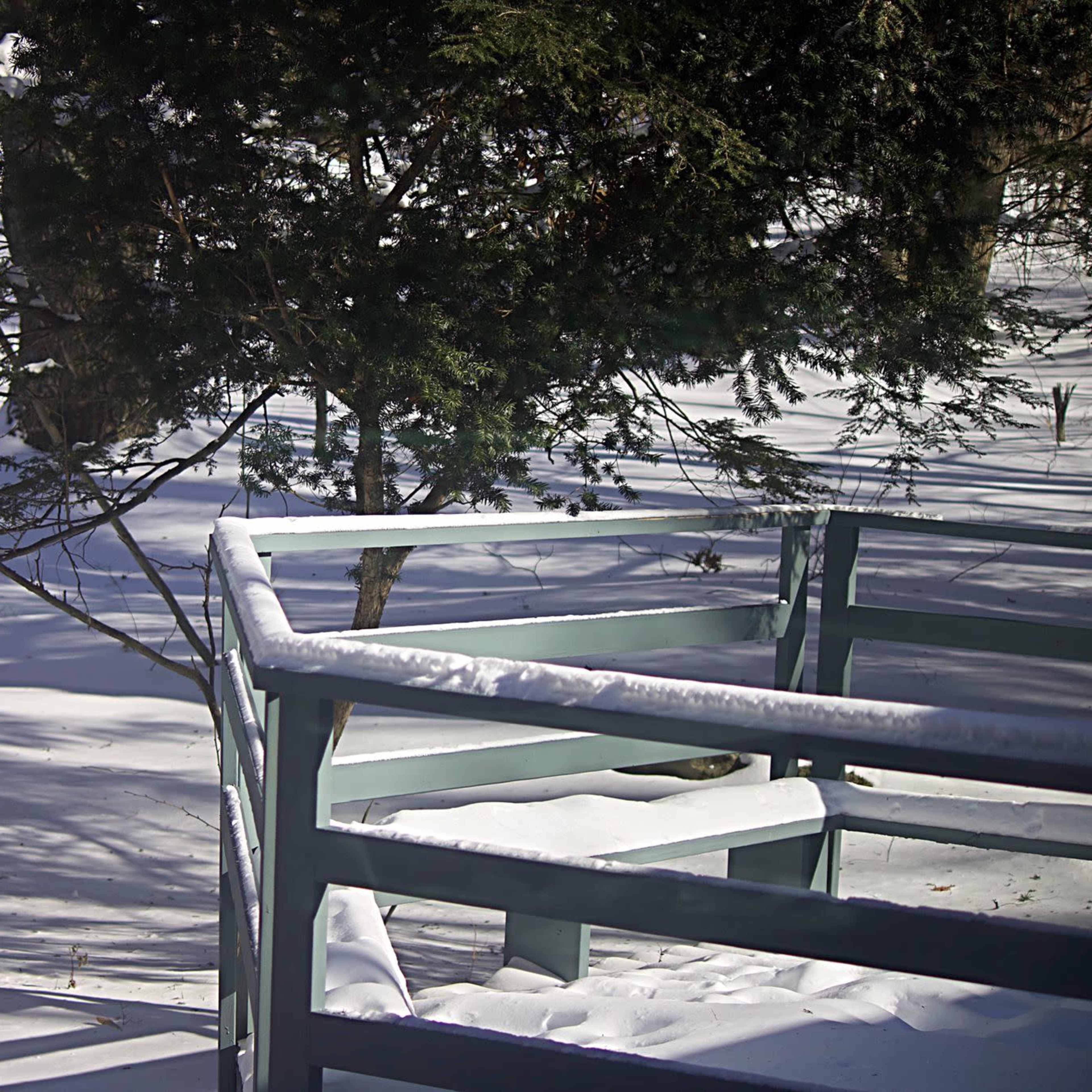 A wooden railing is covered in snow, with trees casting shadows on the snow-covered ground in a winter landscape.