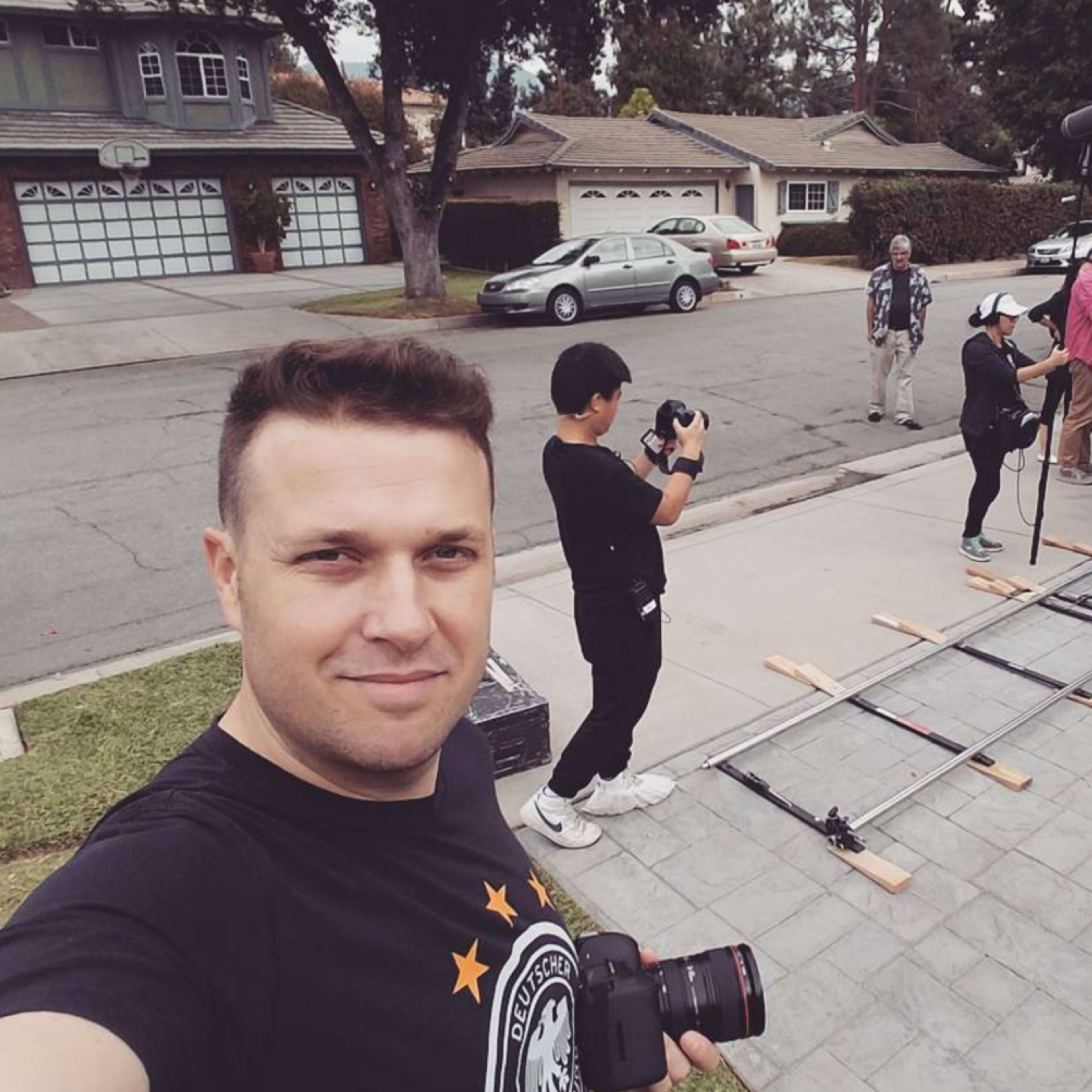 A film crew sets up equipment on a driveway, with one person taking a selfie and others operating cameras and gear.