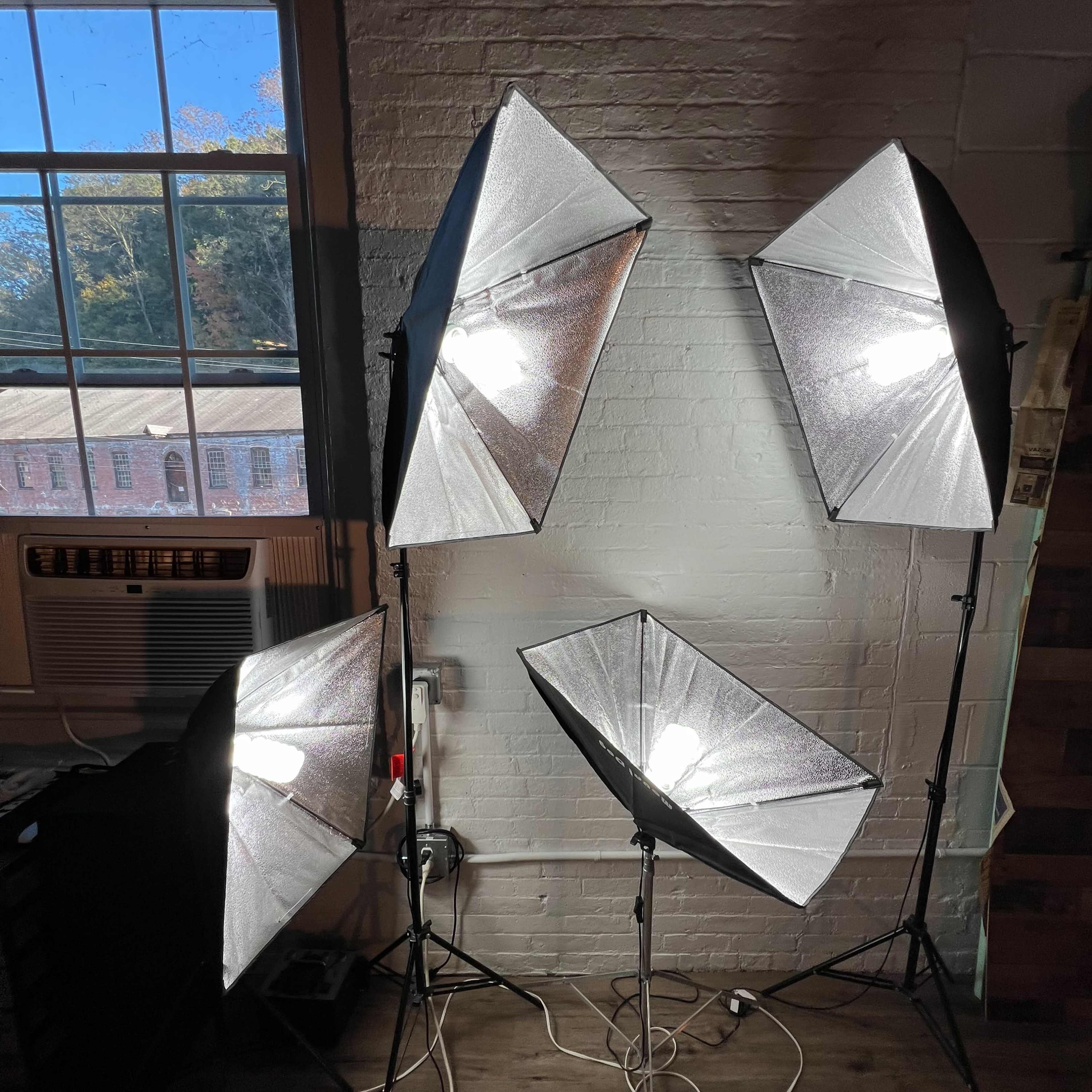 Four softbox lighting setups arranged in a well-lit room with a window and an air conditioning unit in the background.