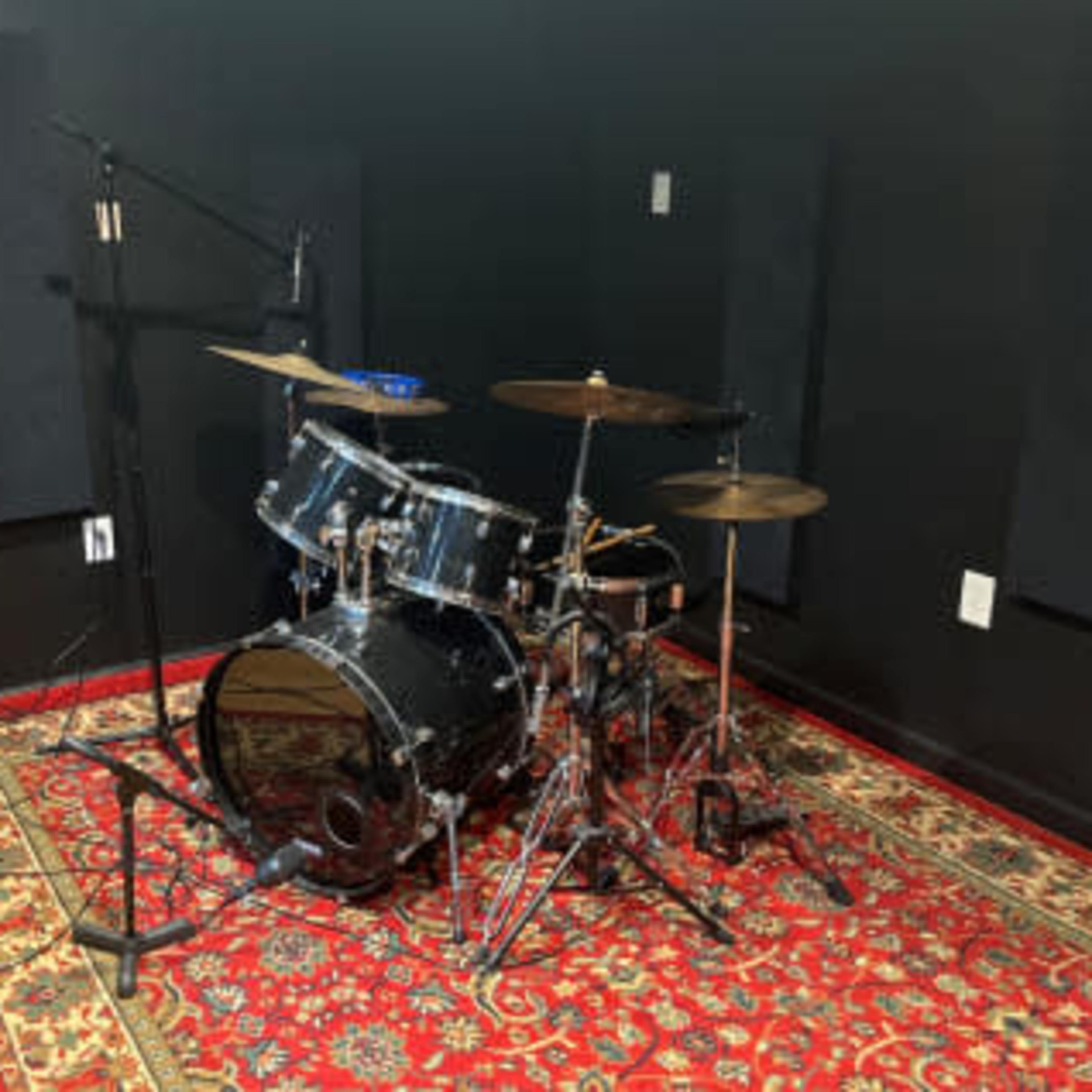 A drum kit is set up on a patterned rug in a soundproofed rehearsal room with black walls and acoustic panels.