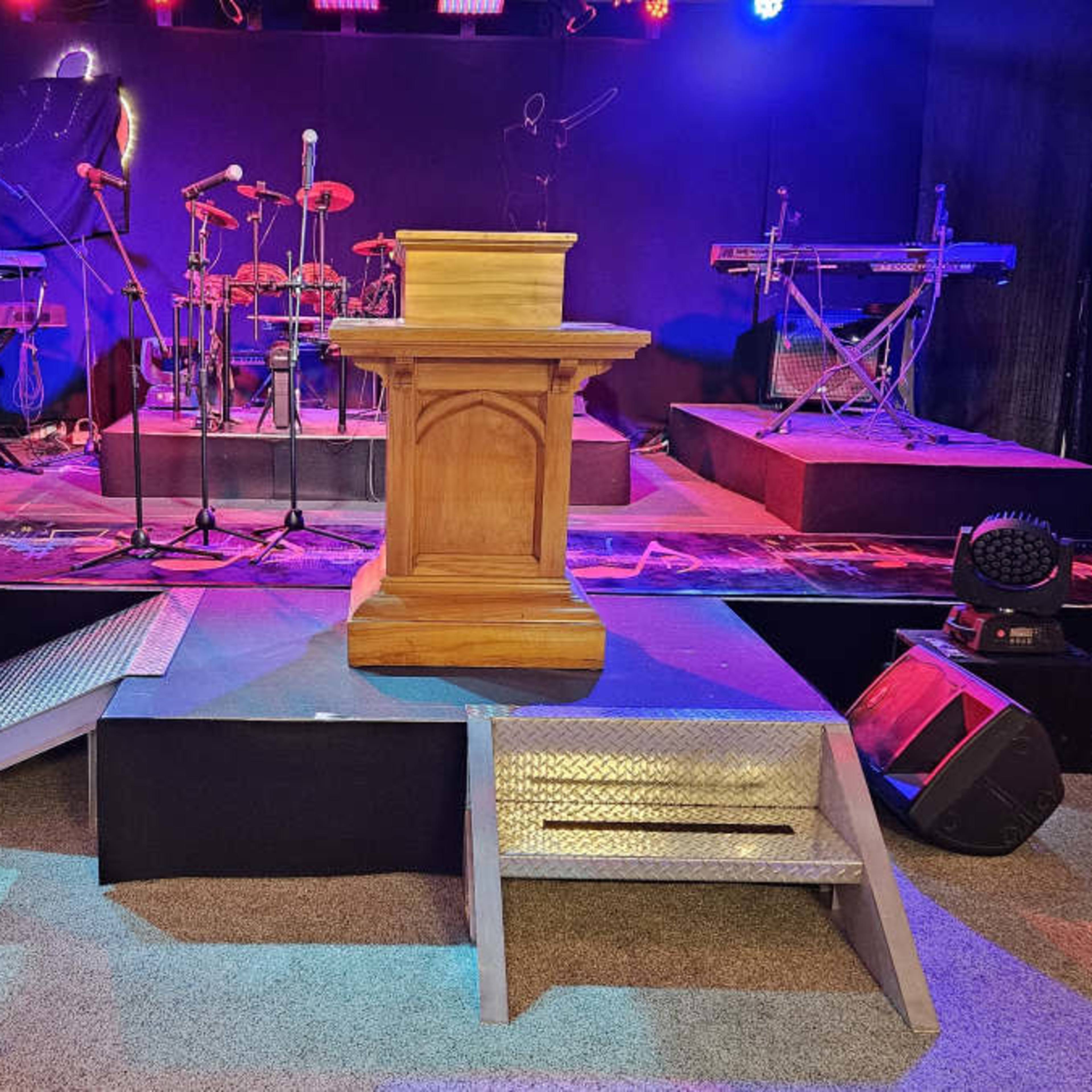 A stage setup with a wooden podium at the center, flanked by musical instruments and equipment under colorful stage lighting.