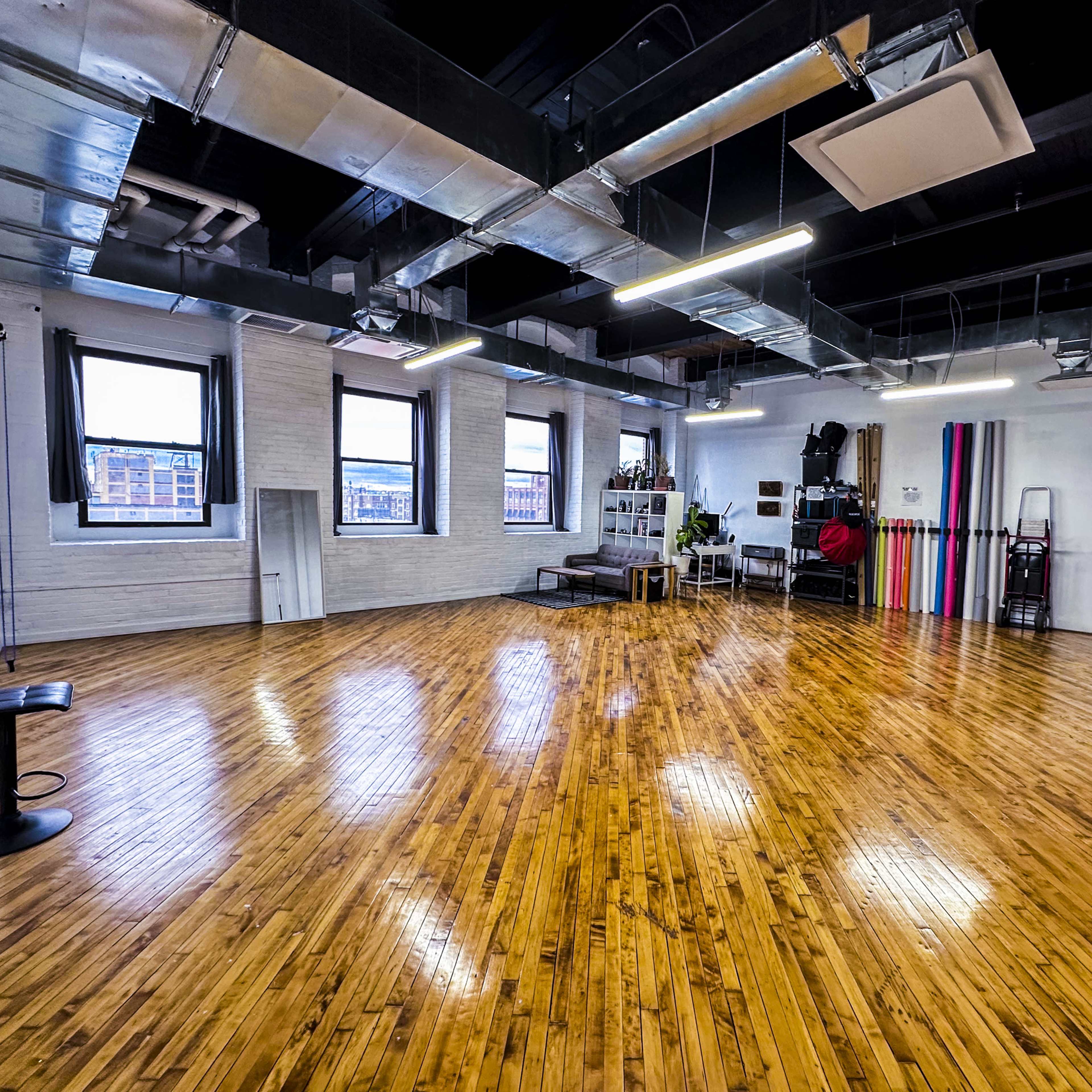 A spacious, empty studio with polished wooden floors, large windows, and various equipment along the walls.
