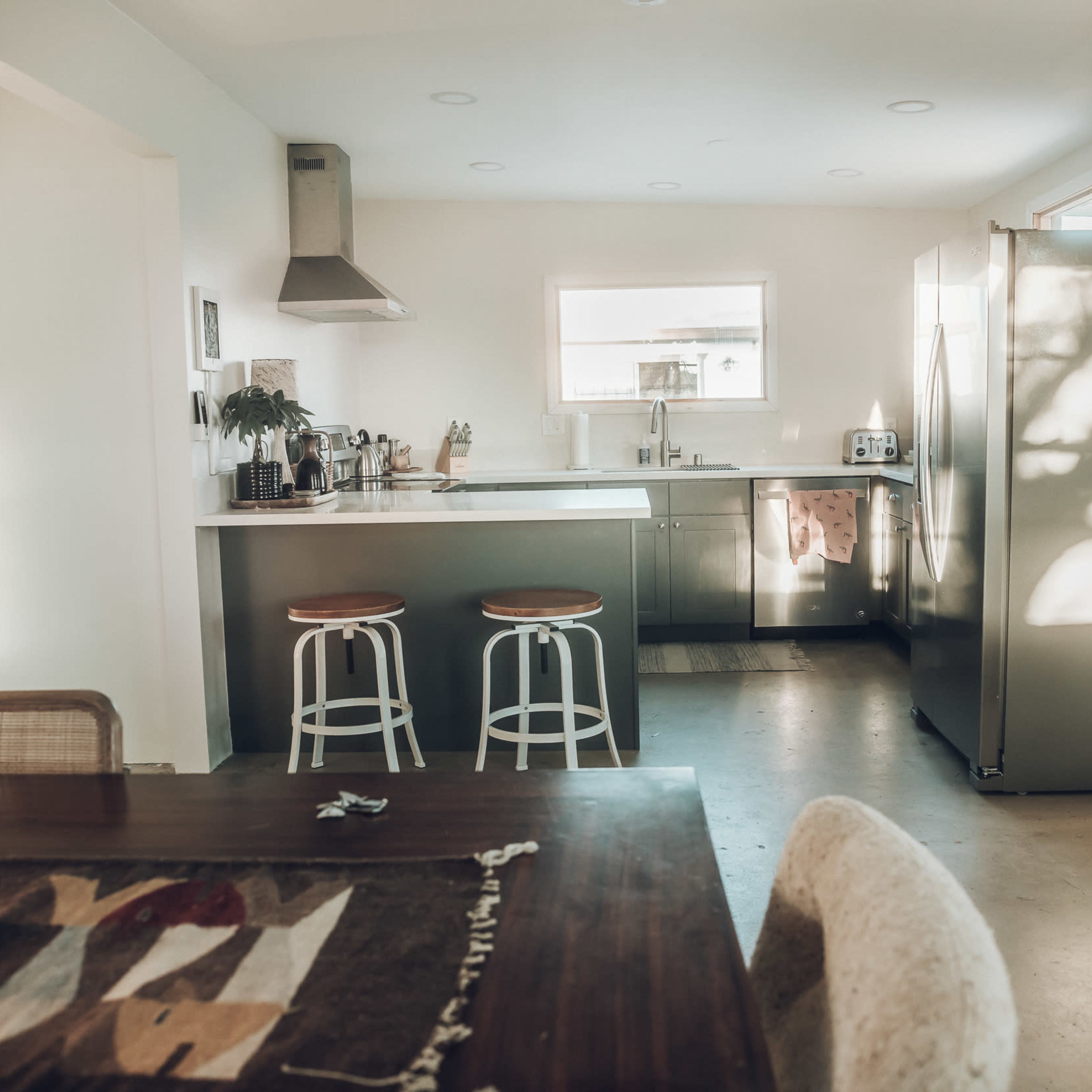 A modern kitchen features stainless steel appliances, a central island with two stools, and a dining table with a decorative tablecloth.