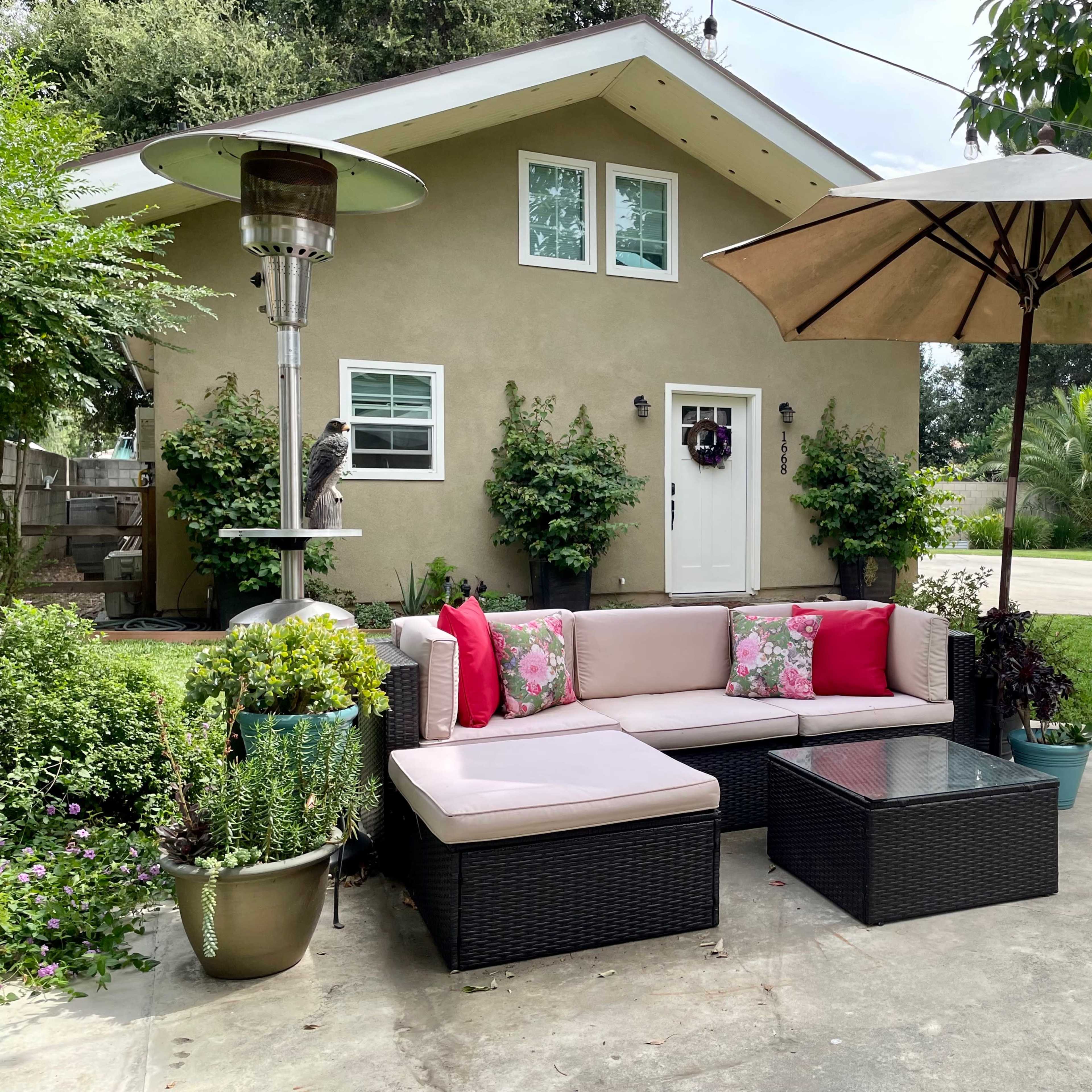 An outdoor seating area with a sectional sofa adorned with pink pillows, a patio umbrella, a gas heater, and a variety of potted plants, all set in front of a small house with a landscaped yard.