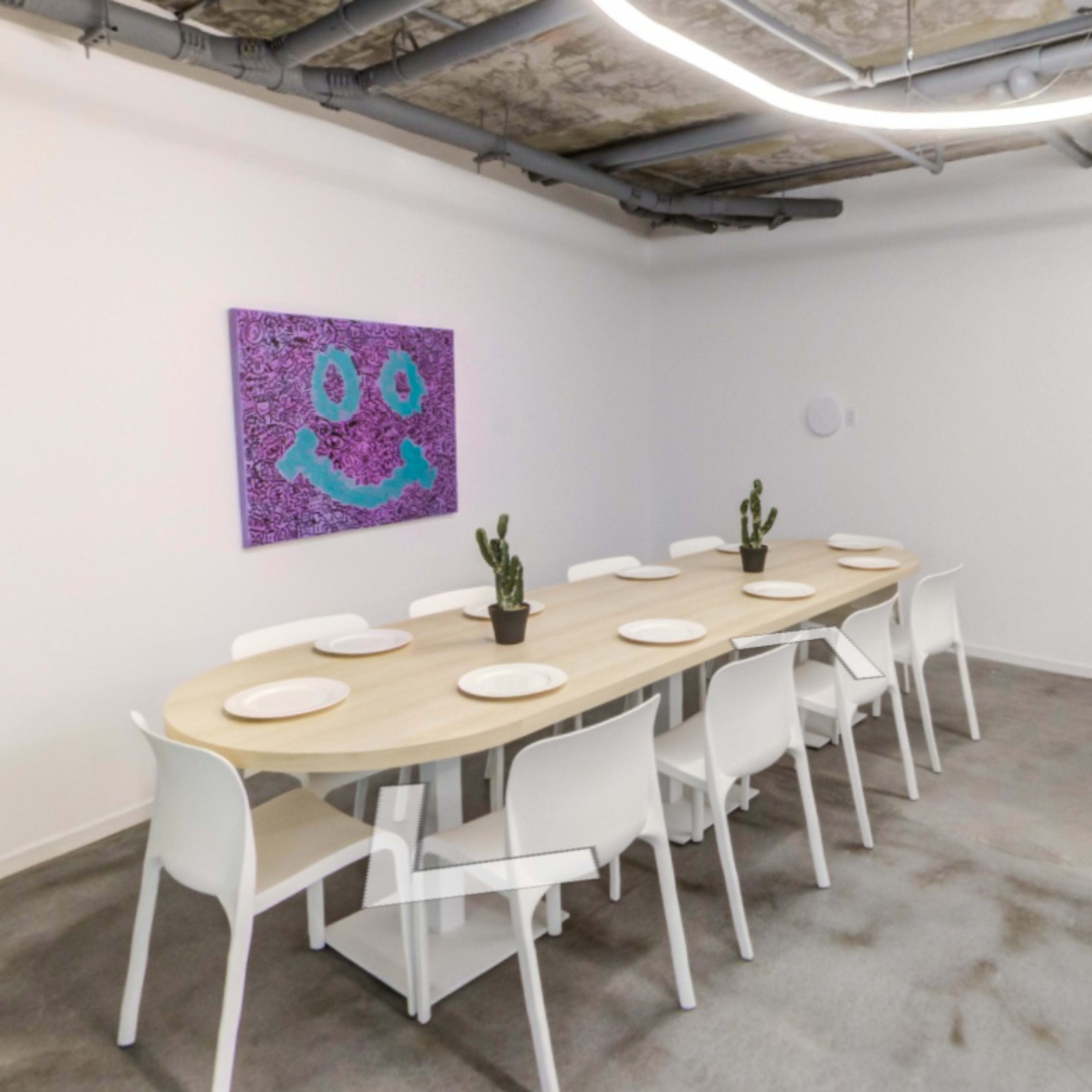 A modern dining area features a long, curved wooden table surrounded by white chairs, with a cactus centerpiece and a smiling face artwork on the wall.