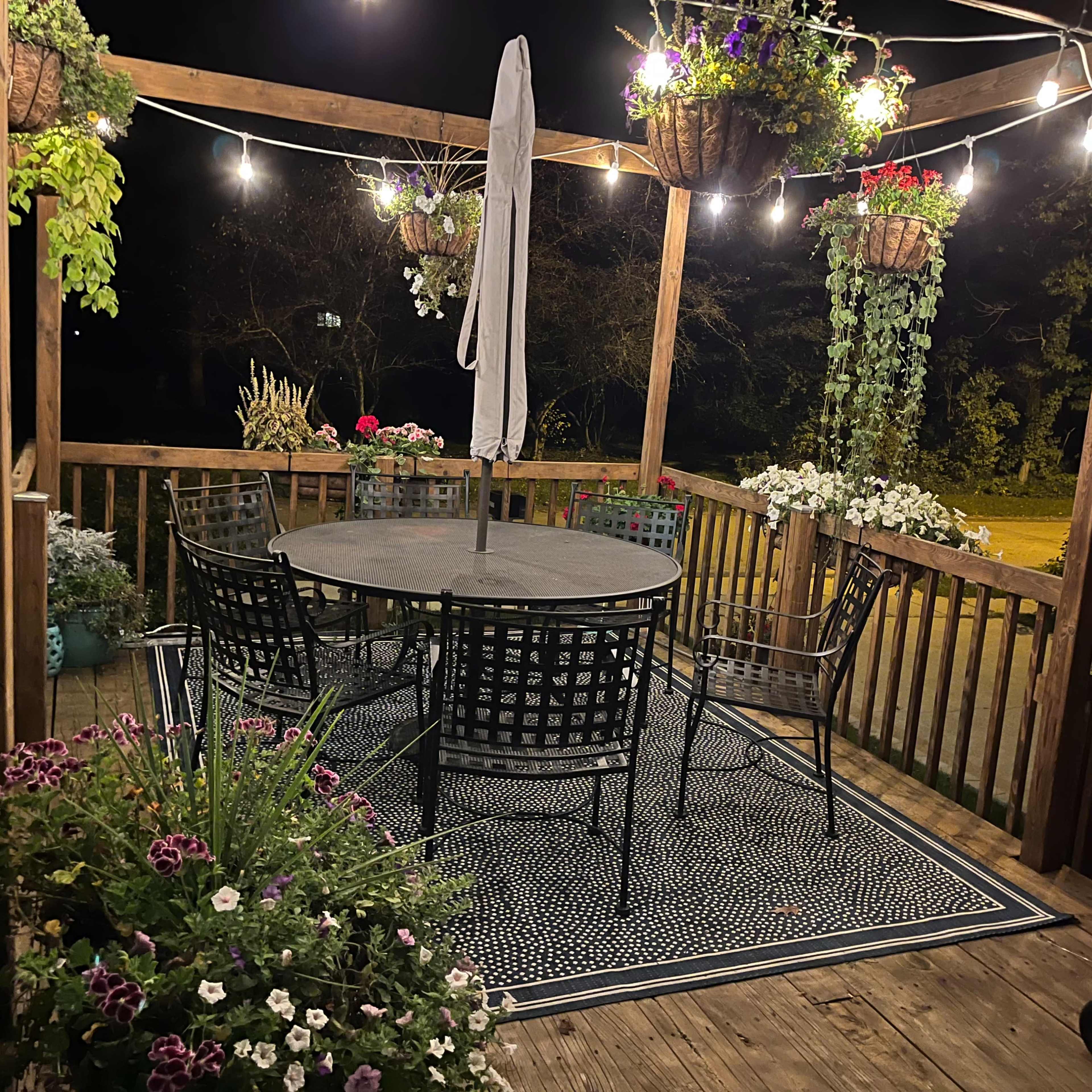 A wooden deck at night features a circular table surrounded by six black chairs, adorned with hanging planters and string lights.