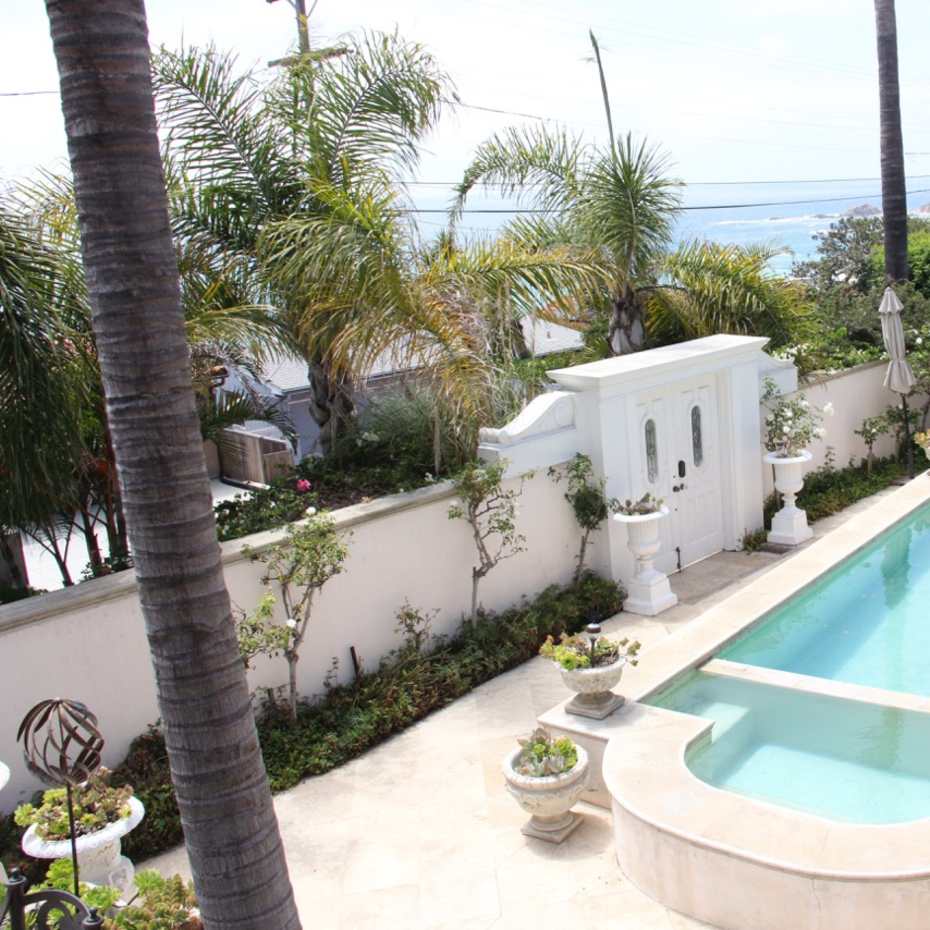 A backyard pool surrounded by tropical plants and seating areas, with a view of the ocean in the background.