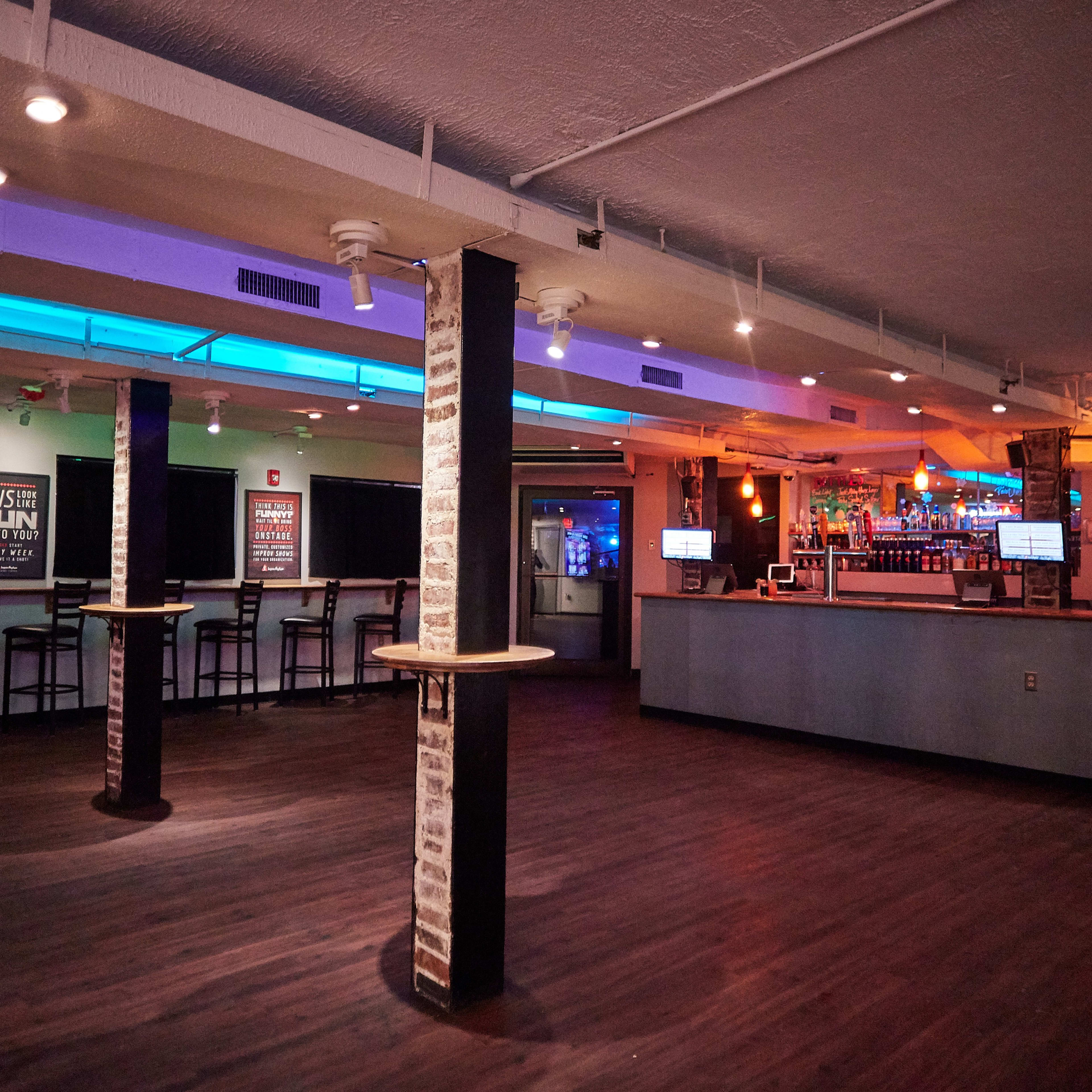An empty bar interior with a counter on the right and rows of chairs along the left wall beneath dim lighting.