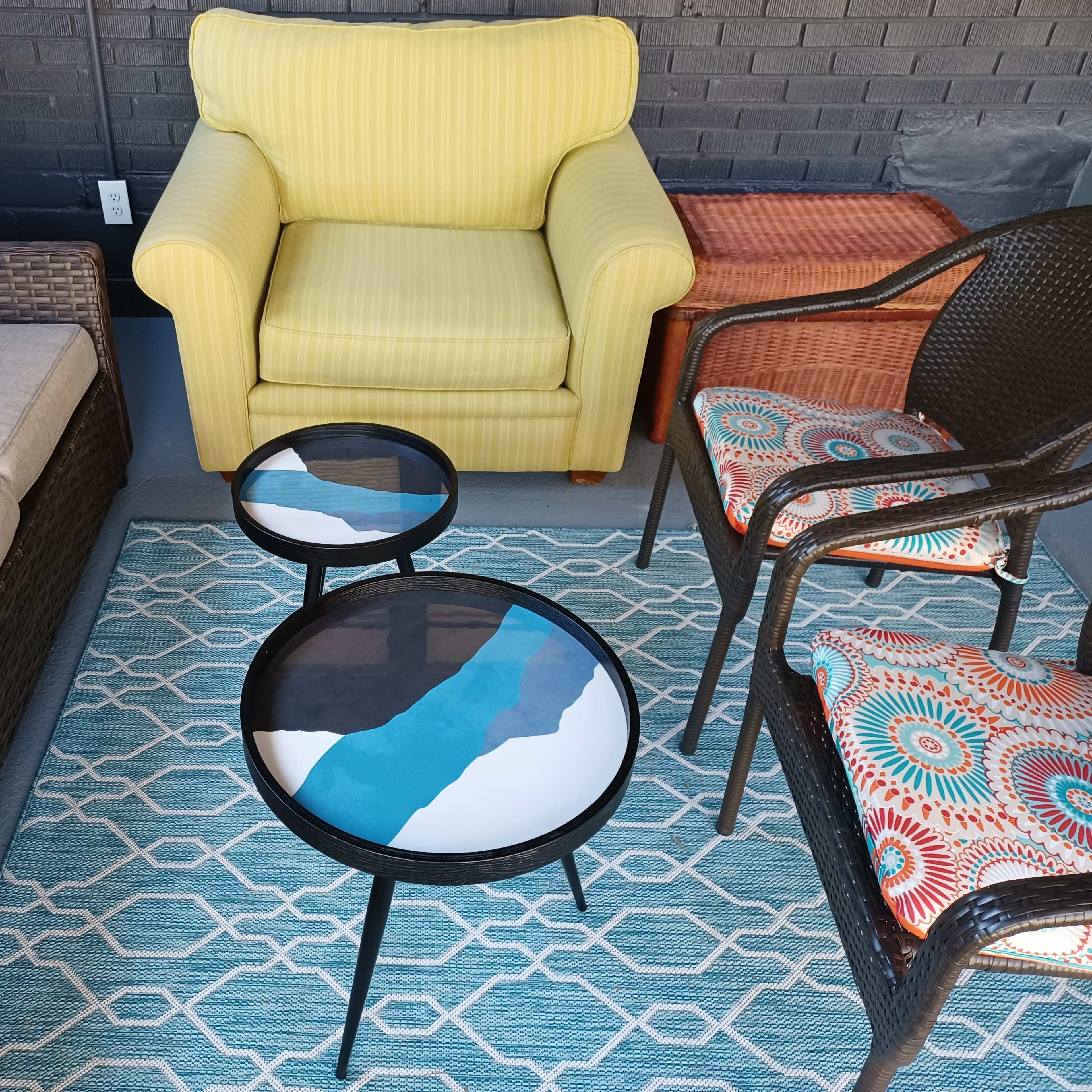 A small outdoor seating area with a yellow armchair, a gray sofa, two round coffee tables, and patterned chair cushions on a geometric rug.