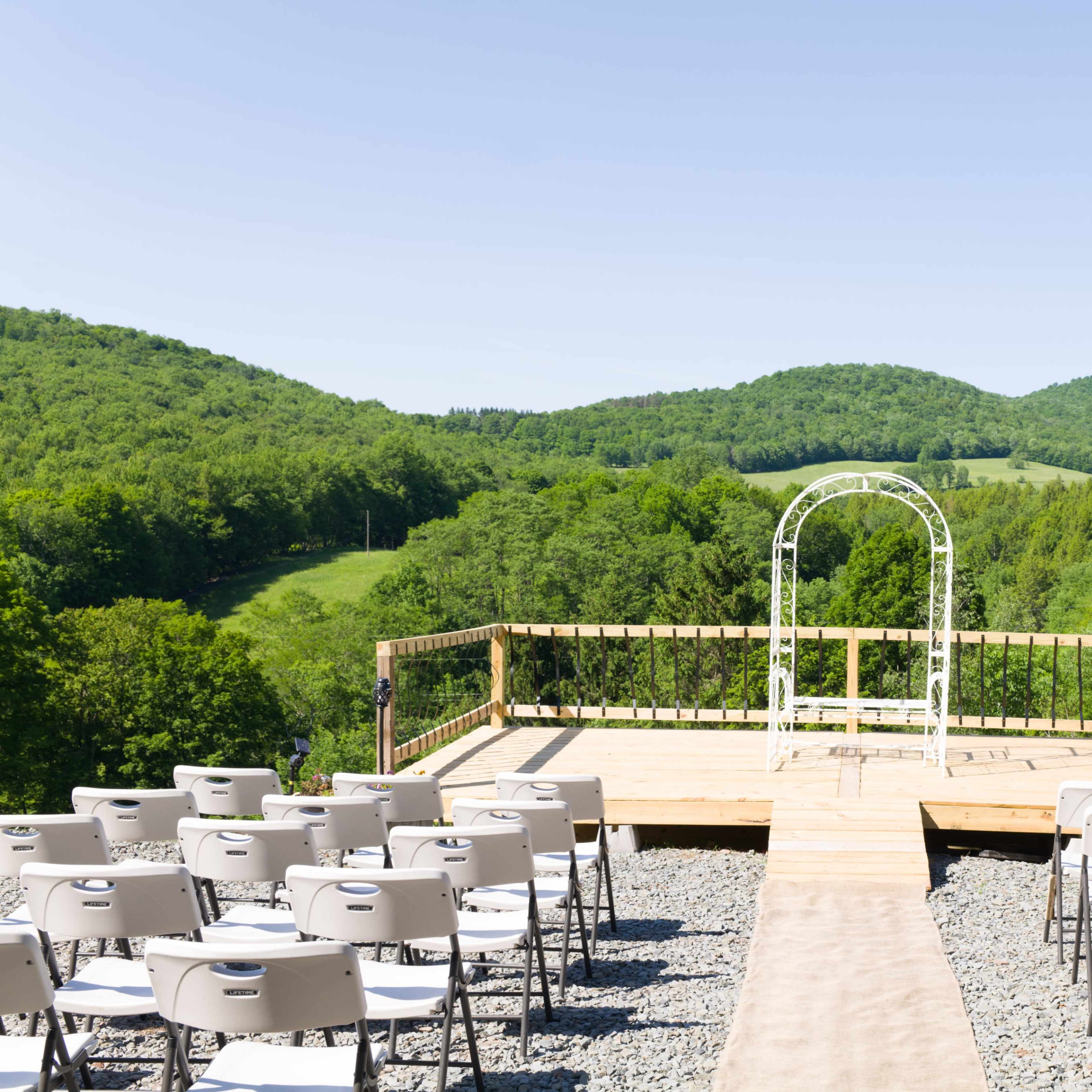 A wooden wedding altar is set up on a platform overlooking a green mountainous landscape, with rows of white chairs facing the altar.