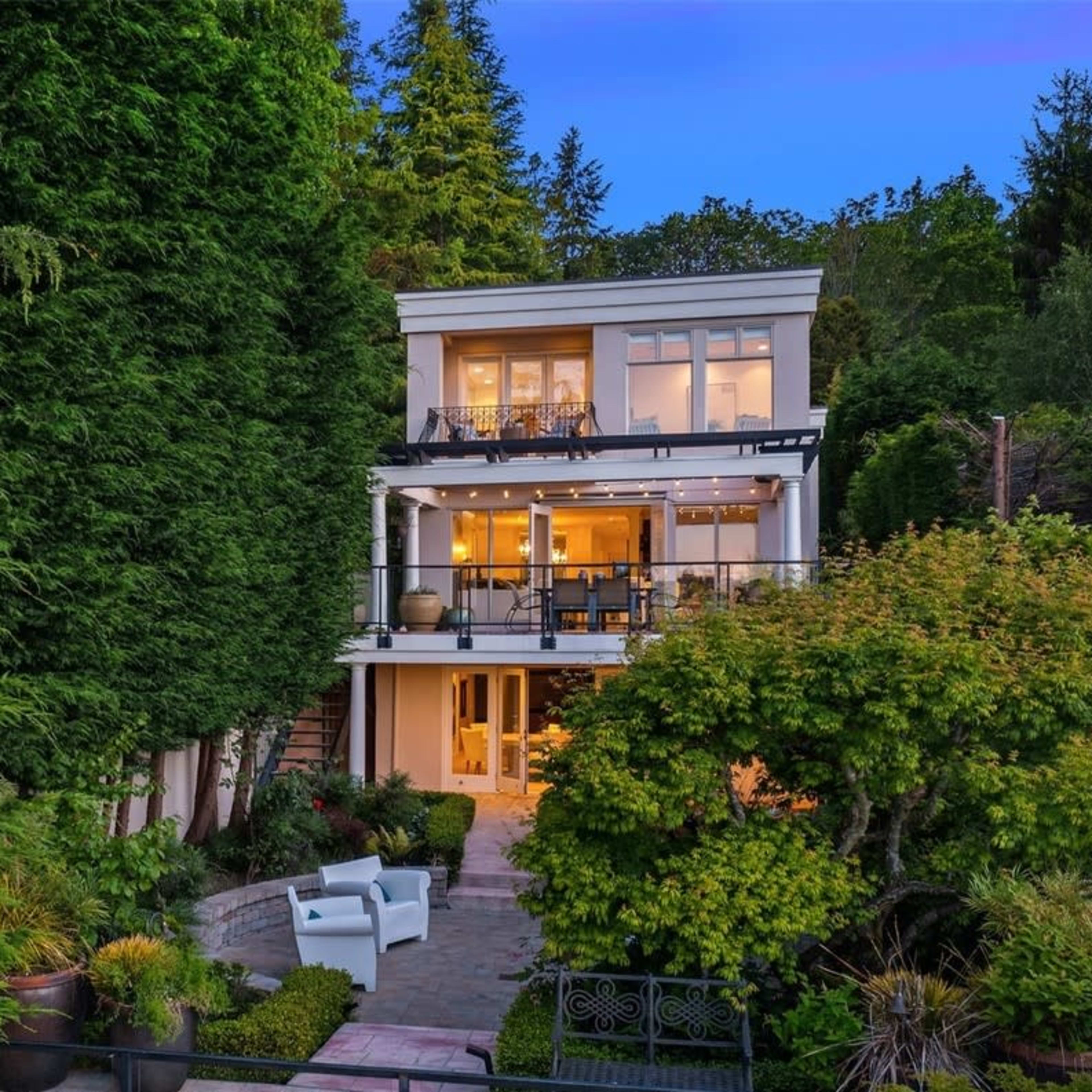 A multi-story house is surrounded by greenery, featuring large windows and a patio area in the front.