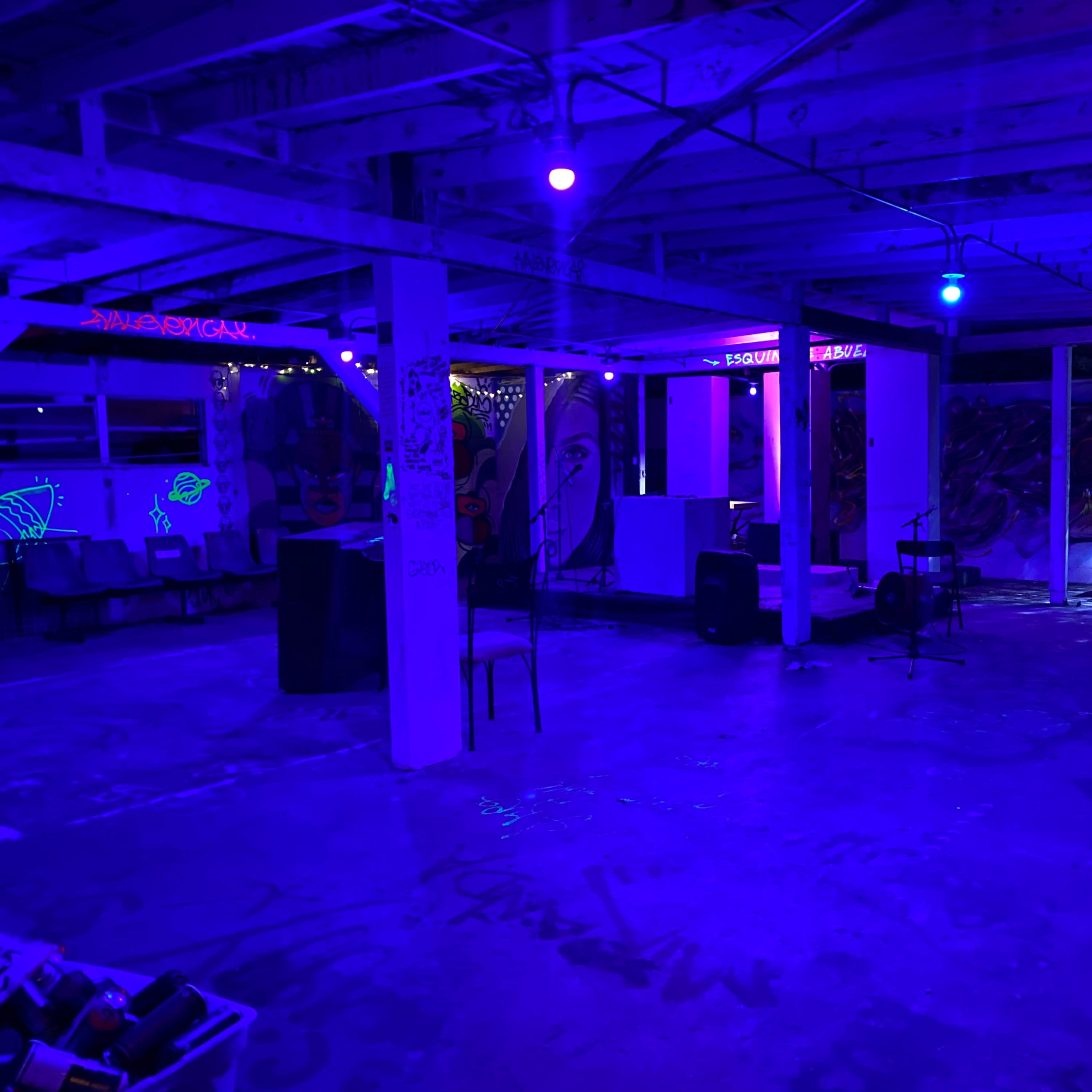 An empty basement space with blue lighting, featuring concrete floors, exposed beams, and scattered chairs and musical equipment.