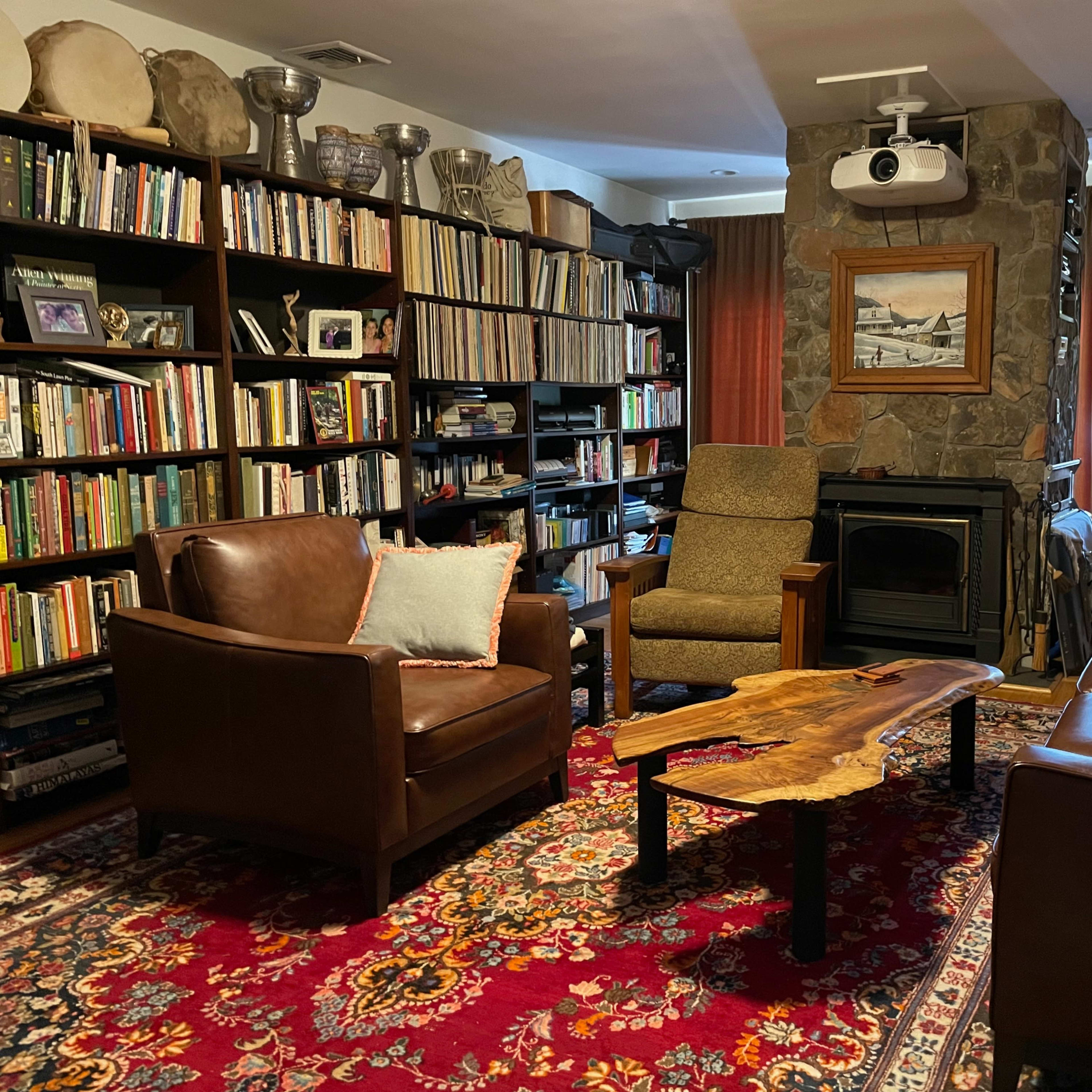 A cozy living room with two leather chairs, a wooden coffee table, and a stone fireplace, surrounded by bookshelves filled with books.