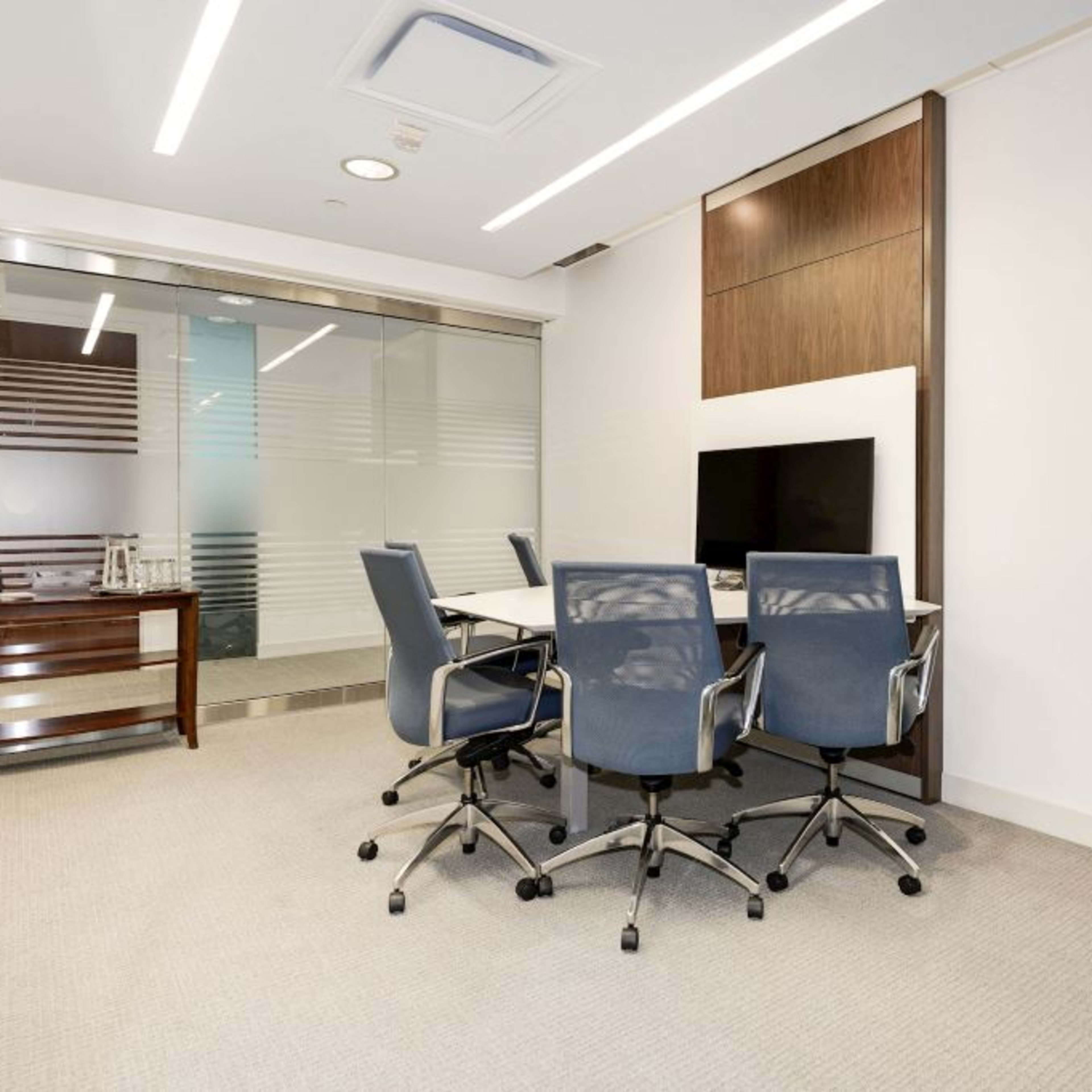 A modern conference room features a glass wall, a rectangular table with four blue chairs, and a kitchenette in the background.