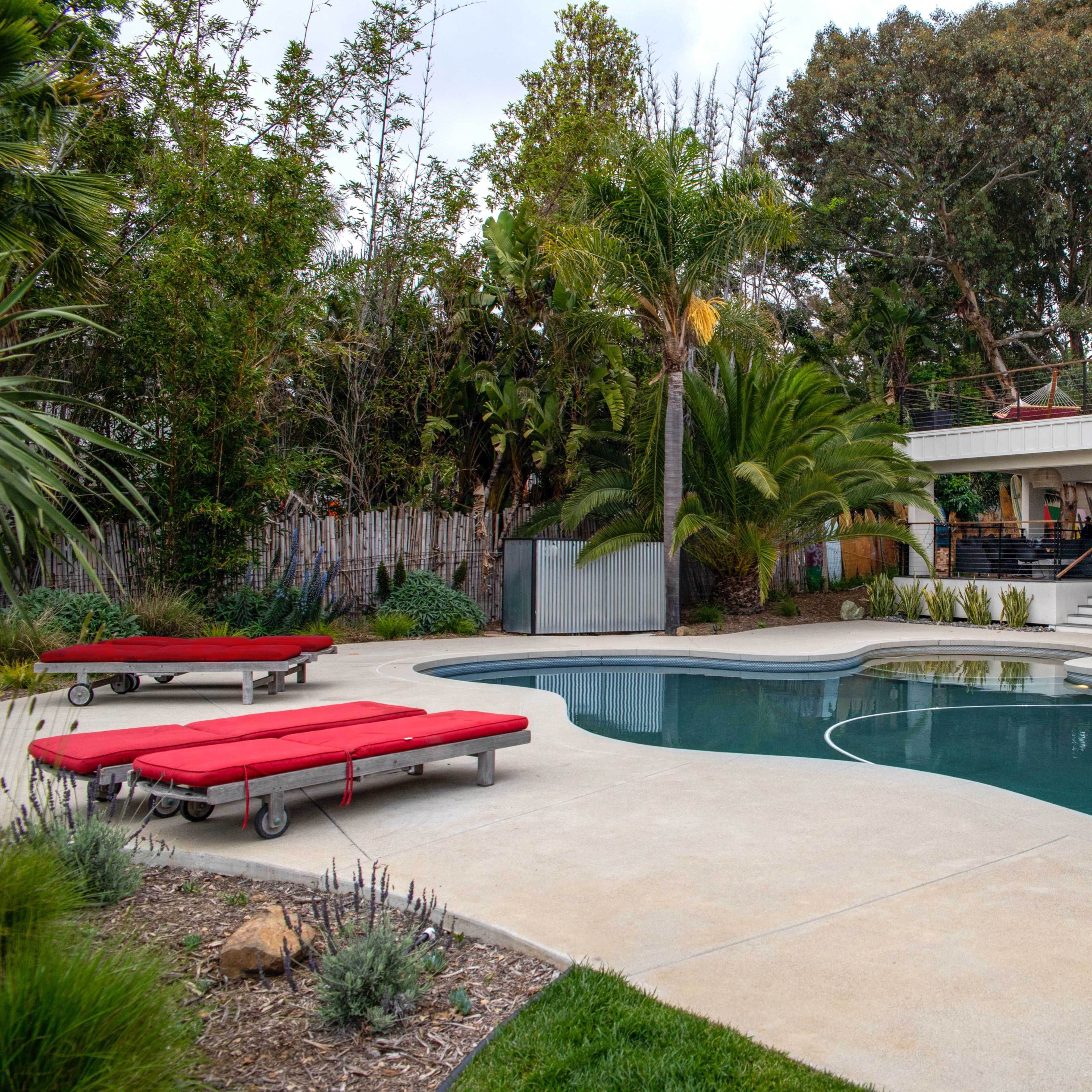 A modern backyard with a swimming pool, two red lounge chairs, and a house featuring a large window and an upper deck.