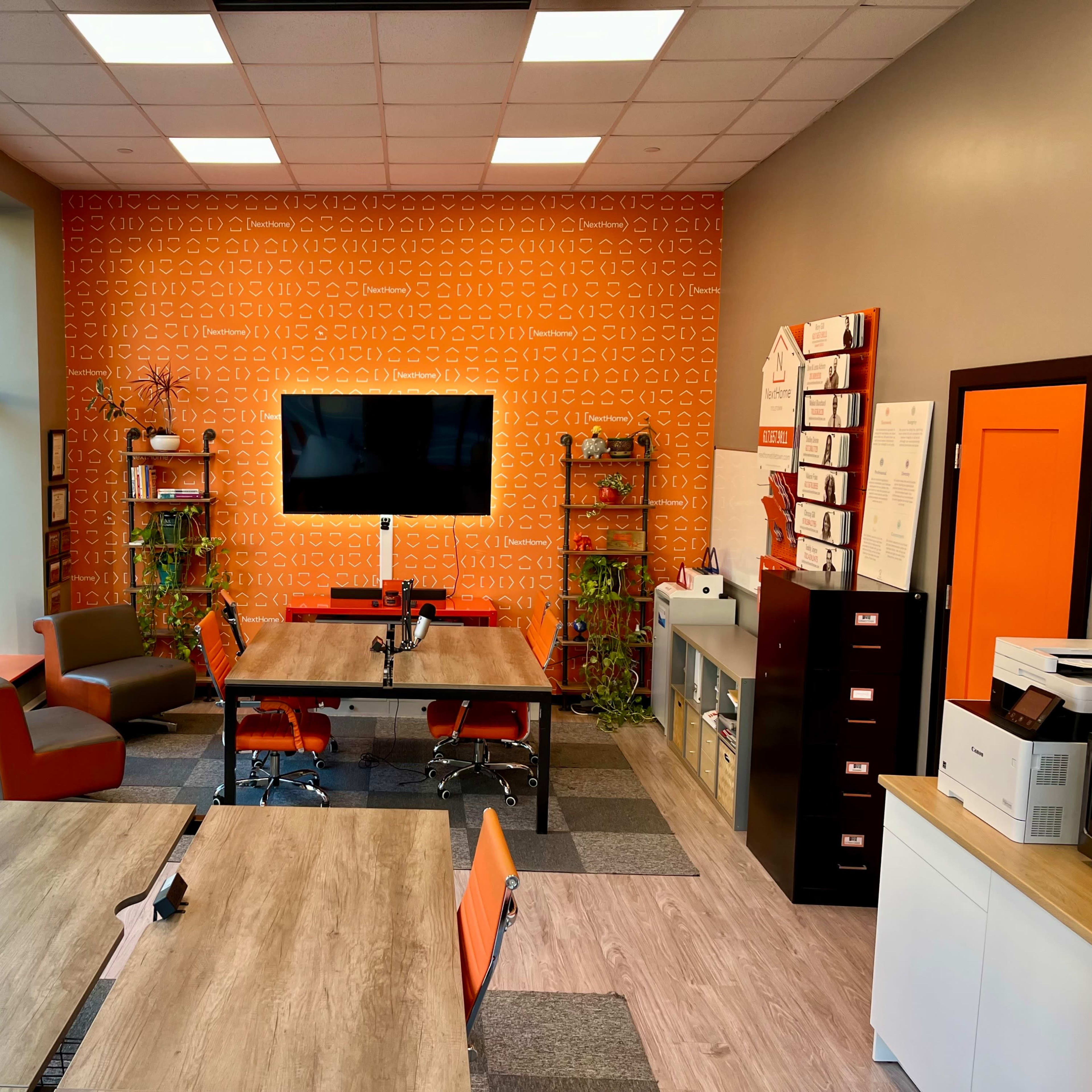A modern office space featuring orange and gray color accents, multiple desks, a large screen on the wall, and green plants.
