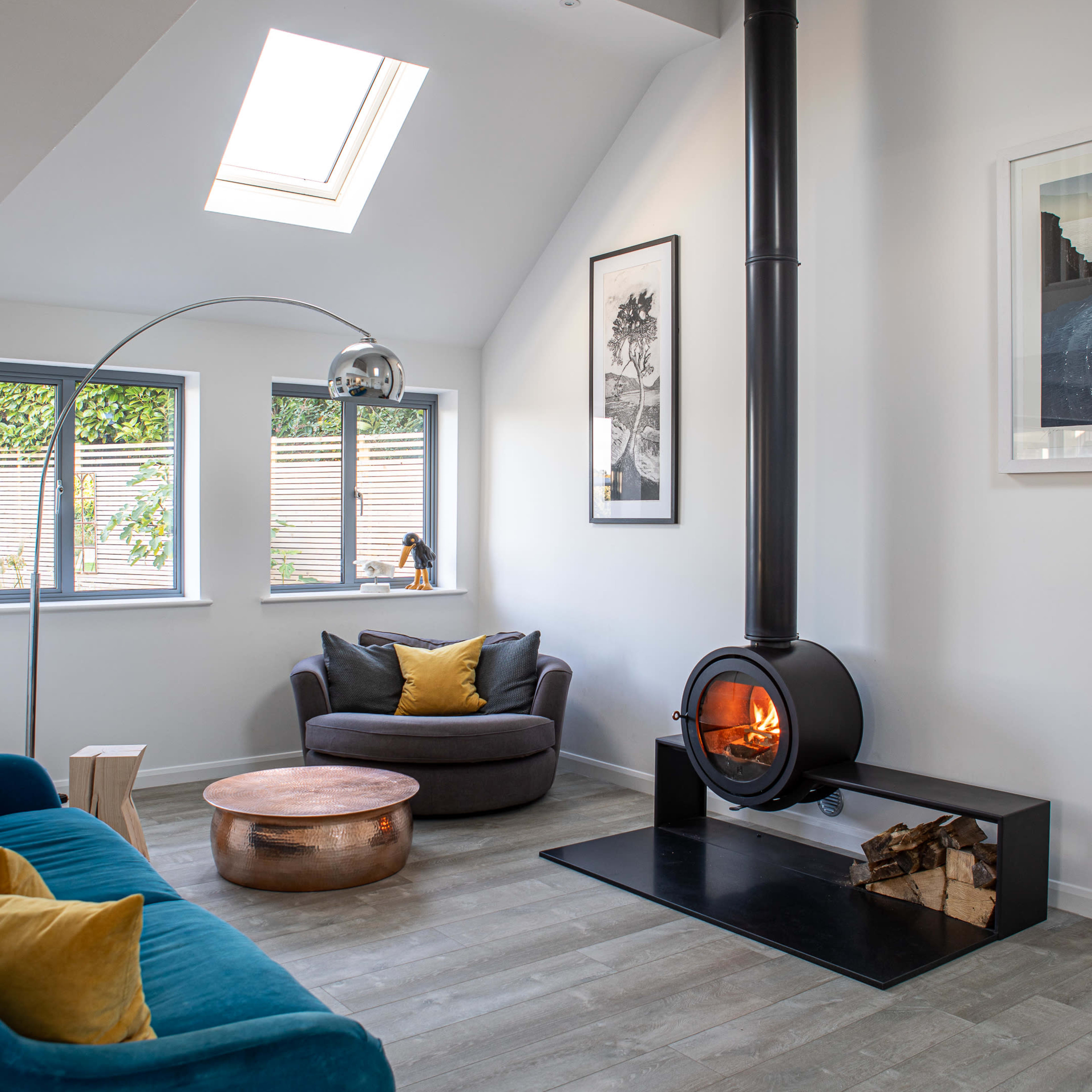 A modern living room features a circular fireplace, a cozy seating area with a blue sofa and a dark armchair, and large windows allowing natural light to enter.