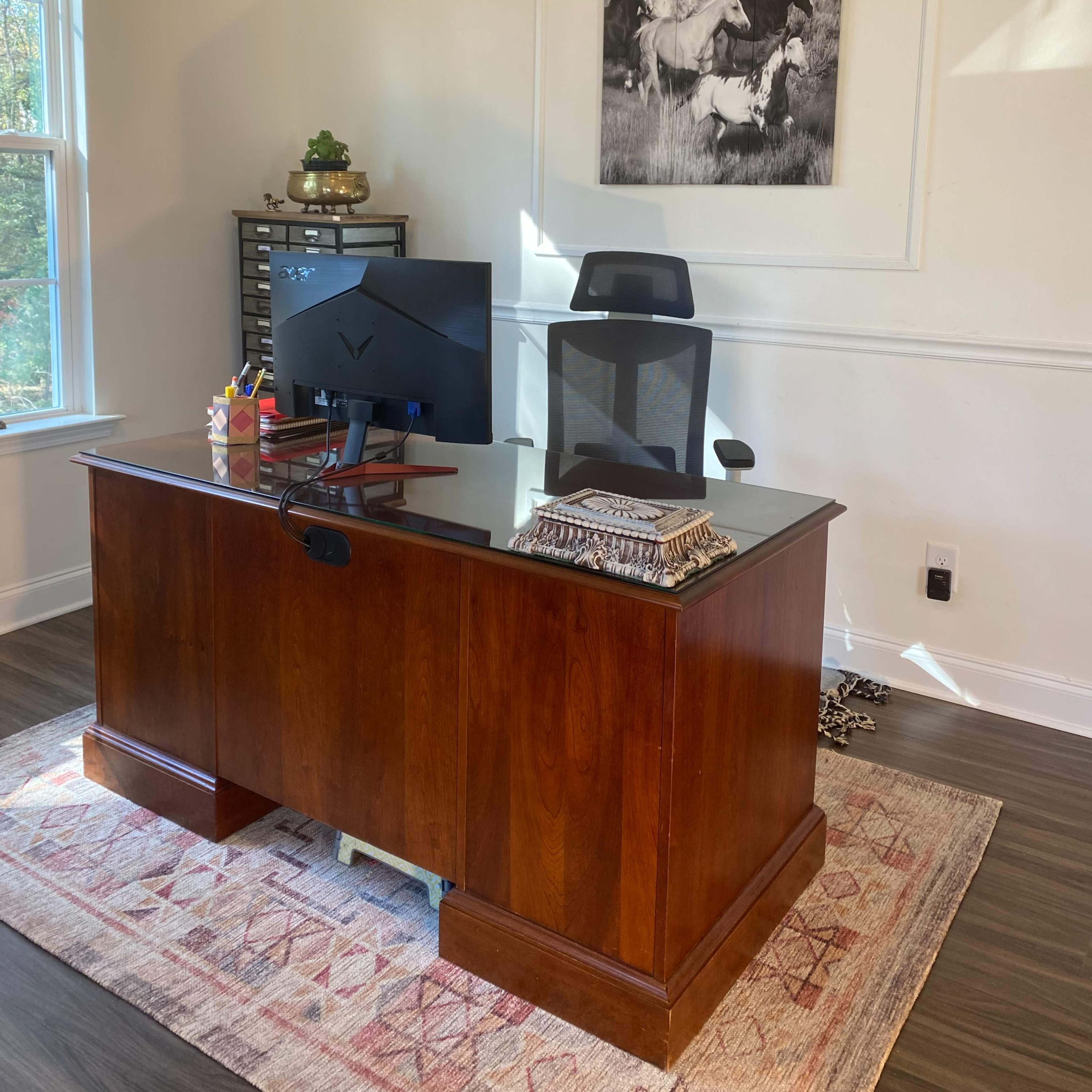 A modern office space featuring a wooden desk with a glass top, a black computer monitor, and a framed picture of horses on the wall.